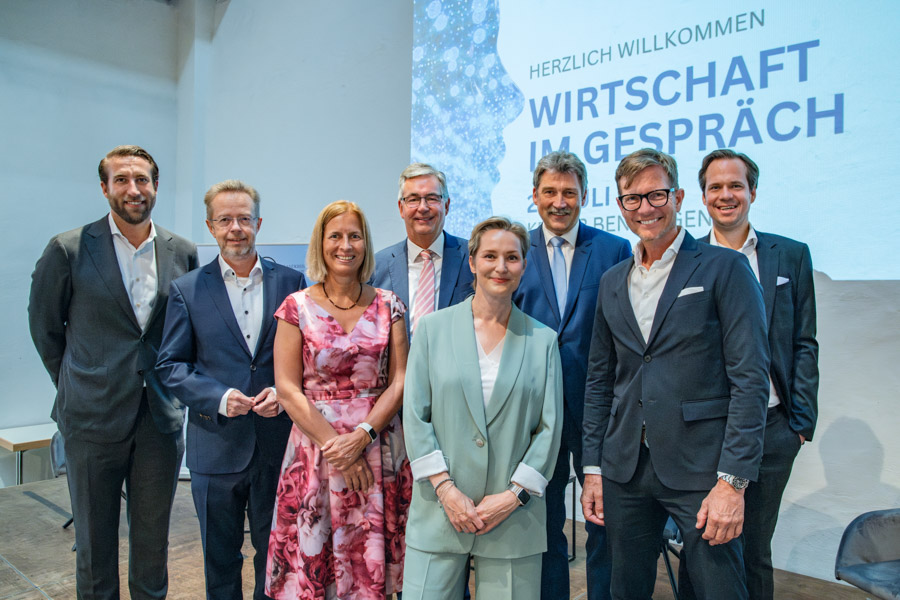 Eine Gruppe von acht Personen steht vor einem Präsentationshintergrund, auf dem „Wirtschaft im Gespräch“ steht. Die Menschen sind formell gekleidet und lächeln in die Kamera. Der Raum hat eine helle, freundliche Atmosphäre.