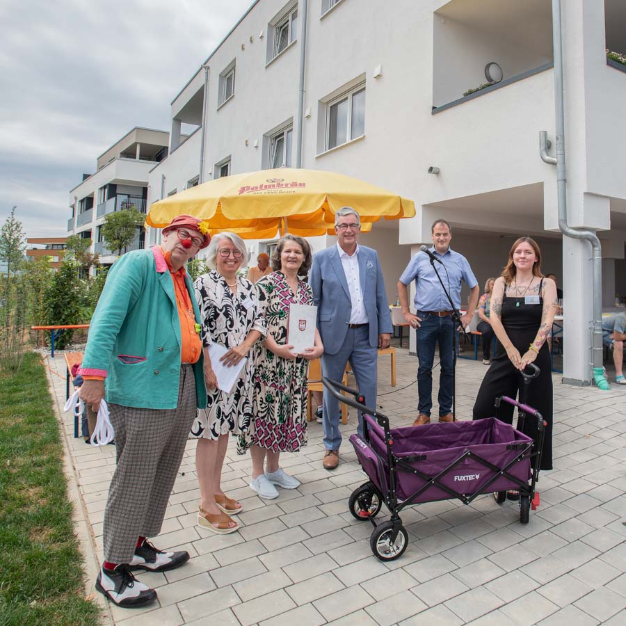 Eine Gruppe von sechs Personen steht im Freien vor einem Gebäude. Eine Person in einem bunten Anzug ist links, während die anderen in formeller Kleidung einen Preis oder eine Auszeichnung halten. Im Hintergrund sieht man eine gelbe Markise.