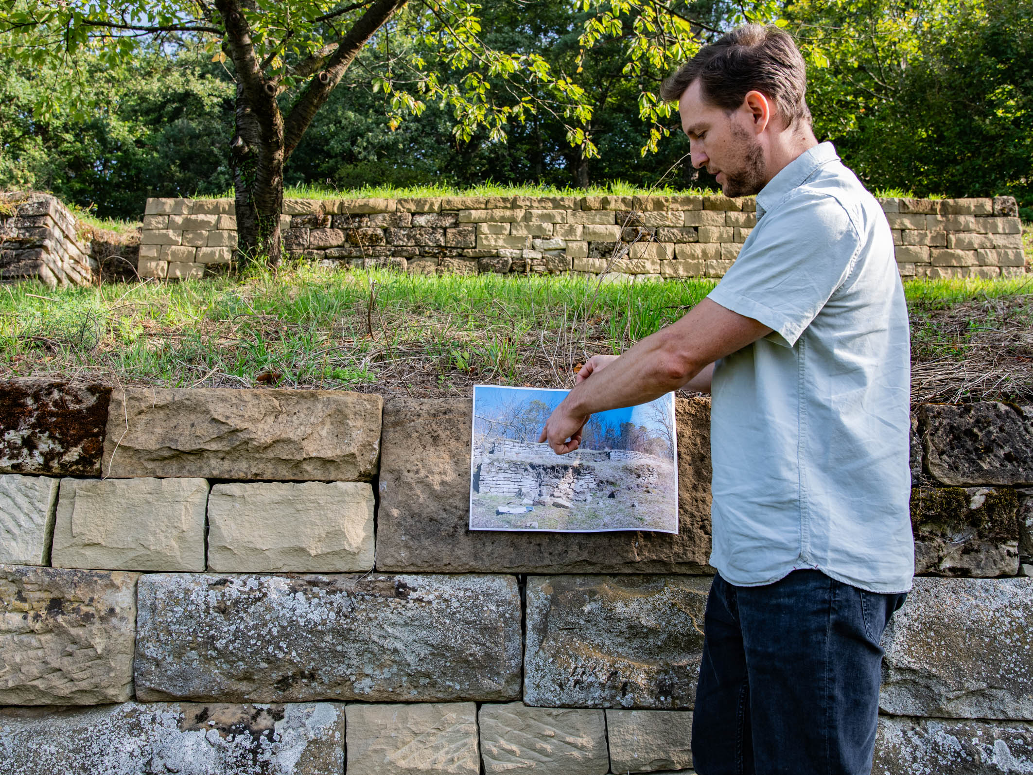 Ein Mann steht an einer Steinmauer und hält ein Bild hoch, das eine alte, zerstörte Landschaft zeigt. Er zeigt mit dem Finger auf einen Bereich des Bildes, während im Hintergrund eine grüne Umgebung mit Bäumen zu sehen ist.