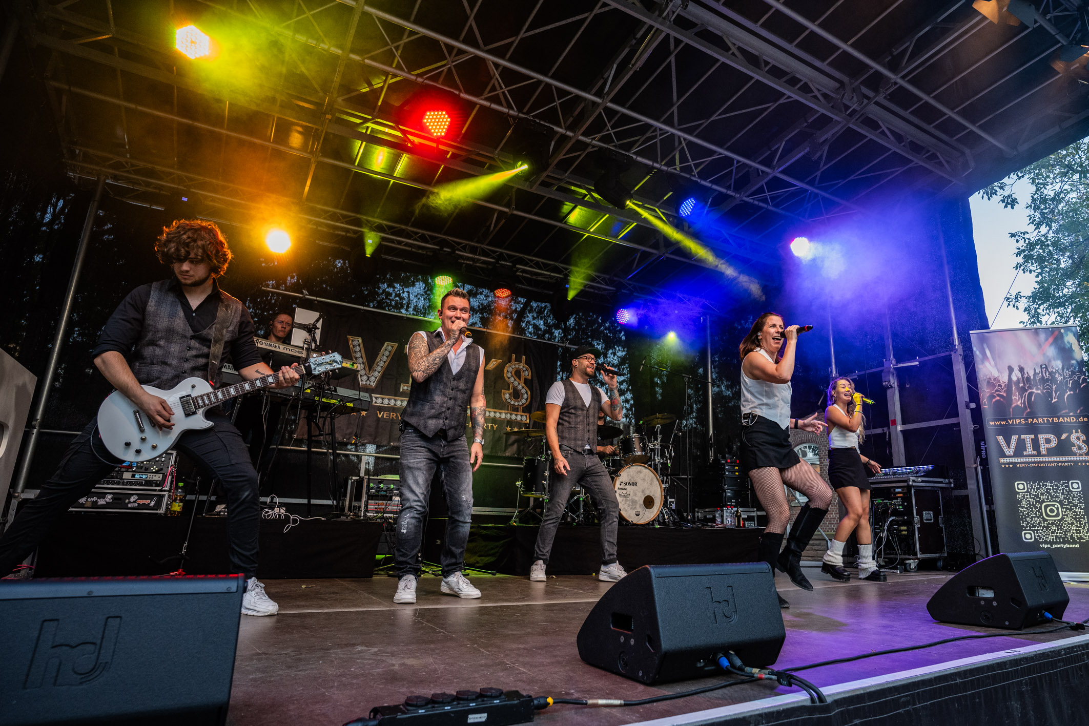 Eine Band führt auf einer Bühne ein Live-Konzert auf. Mitglieder der Band spielen Instrumente und singen, während bunte Lichter die Atmosphäre beleuchten. Die Stimmung ist energetisch und lebhaft.