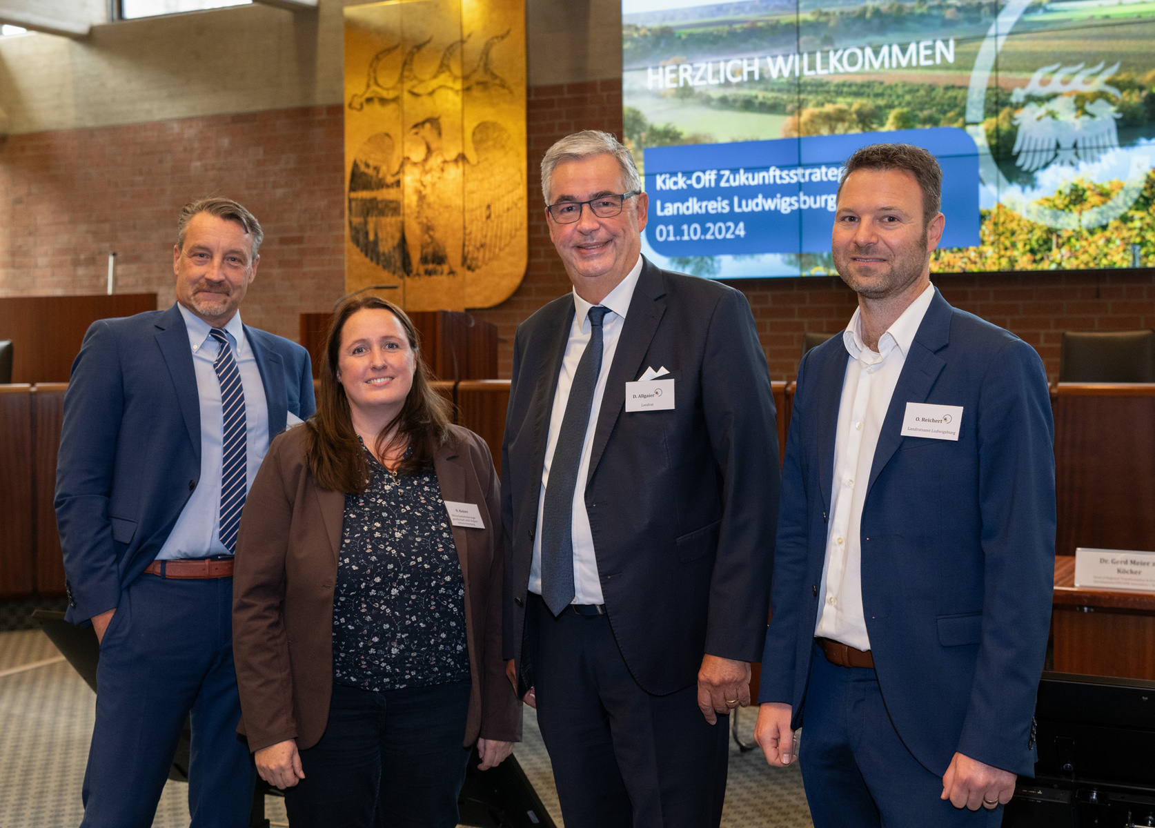 Vier Personen stehen zusammen in einem Raum, der für eine Veranstaltung vorbereitet ist. Im Hintergrund ist ein Bildschirm mit dem Text „Herzlich Willkommen“ und Informationen zu einer Kick-Off-Veranstaltung für die Zukunftsstrategie des Landkreises Ludwigsburg am 01.10.2024.