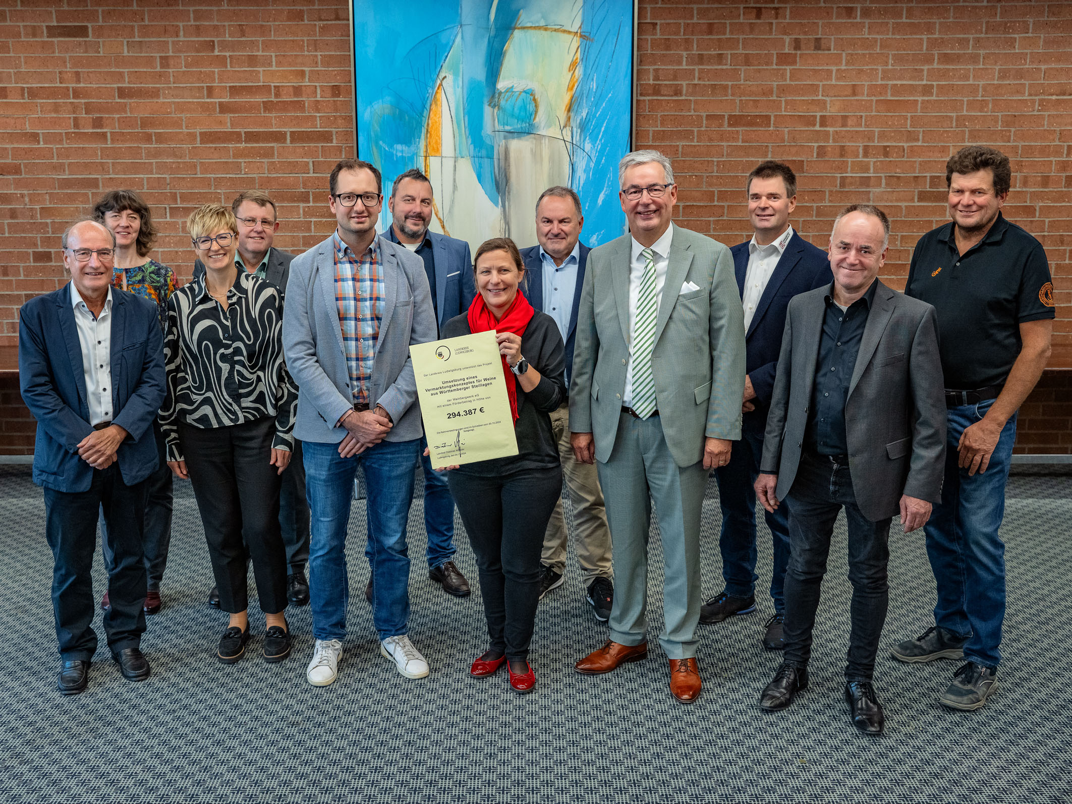 Eine Gruppe von zwölf Personen steht in einem Raum vor einer gemusterten Wand. Sie halten ein Zertifikat in der Mitte. Die Gruppe besteht aus Männern und Frauen unterschiedlichen Alters, die lächeln und in formeller Kleidung gekleidet sind.