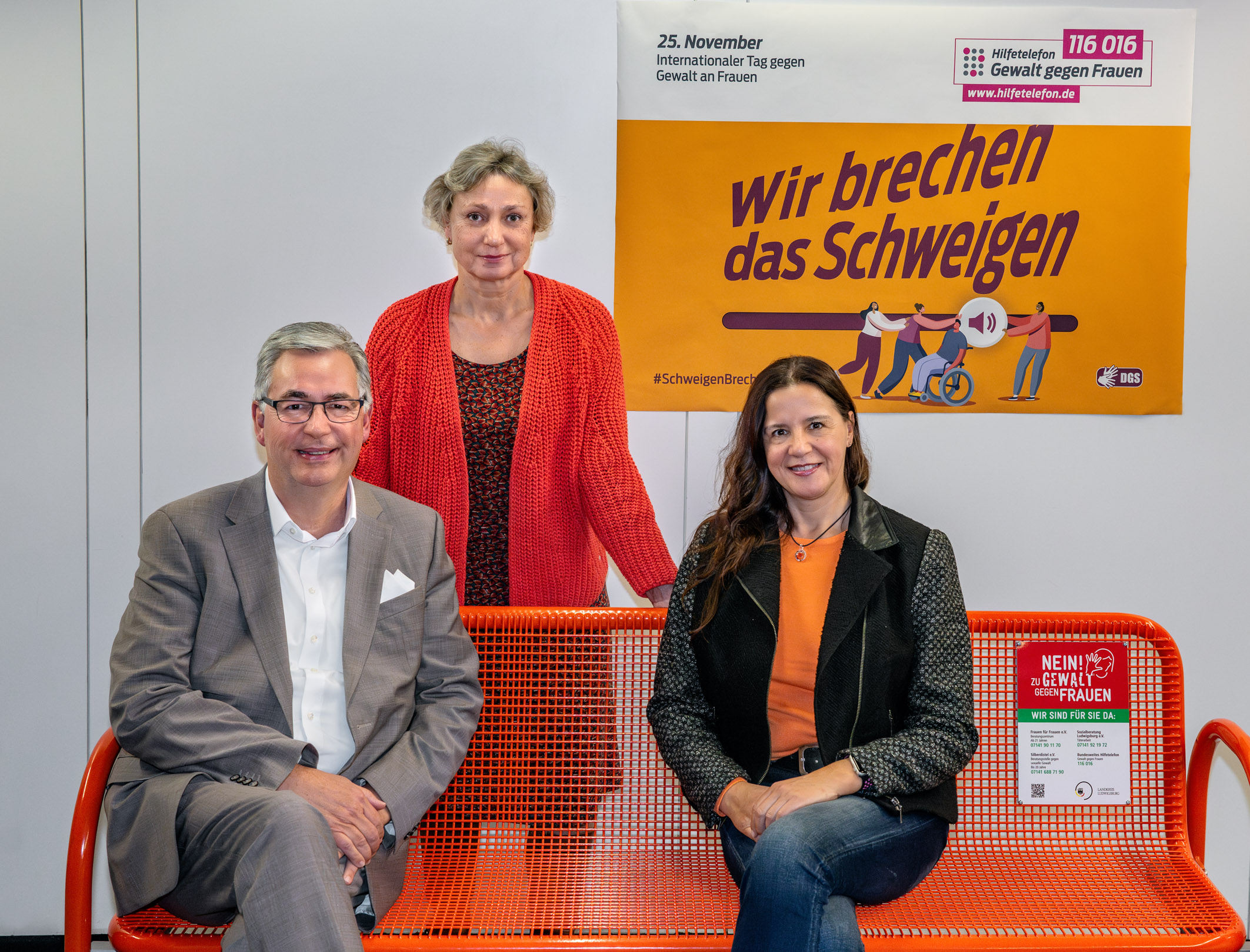 Drei Personen sitzen auf einer orangen Bank in einem Raum mit einem Plakat im Hintergrund. Das Plakat trägt den Text „Wir brechen das Schweigen“ und thematisiert den Internationalen Tag gegen Gewalt an Frauen am 25. November.