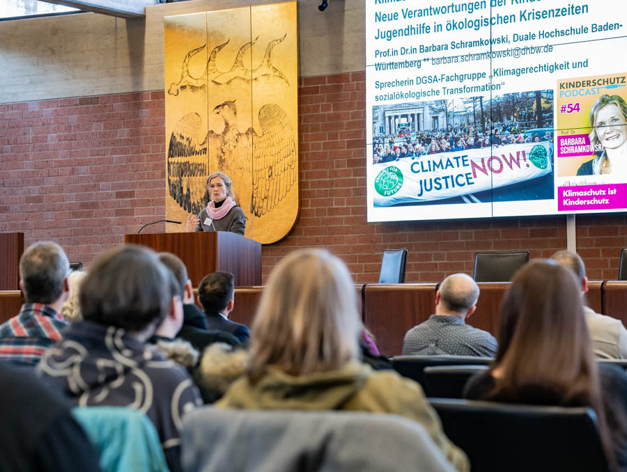 Eine Frau steht an einem Rednerpult und spricht vor einem Publikum in einem großen Saal. Im Hintergrund sind Präsentationsfolien mit dem Titel "Klimagerechtigkeit jetzt!" und Informationen zur ökologischen Transformation zu sehen. Manche Zuhörer wenden sich der Rednerin zu.