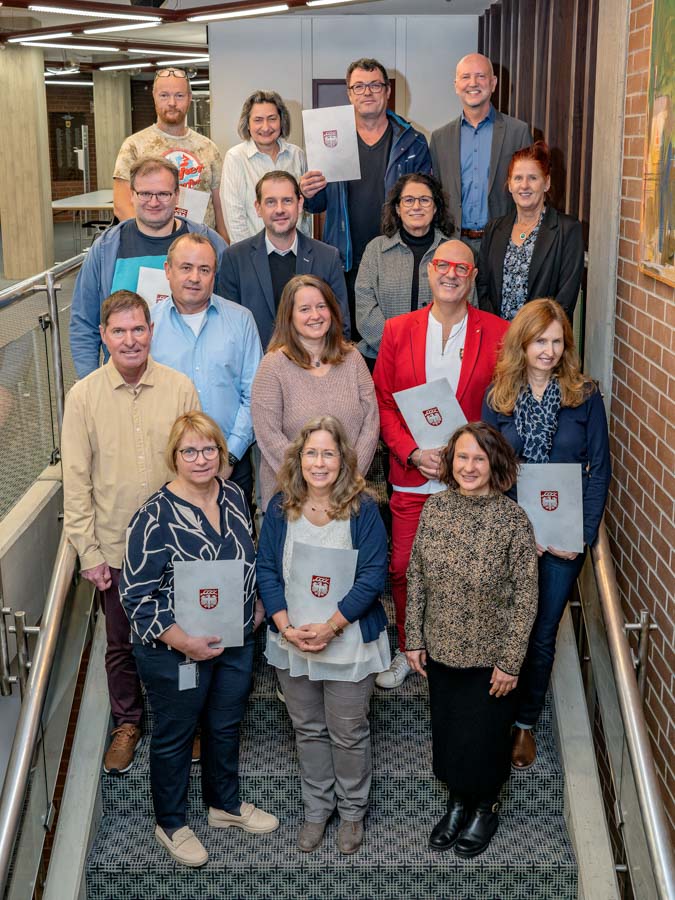 Eine Gruppe von 16 Personen steht auf einer Treppe und hält Dokumente in den Händen. Die Menschen lächeln und tragen verschiedene Kleidung. Im Hintergrund sind Wände aus Ziegelstein und Holz sichtbar. Die Gruppe scheint an einem feierlichen Anlass teilzunehmen.