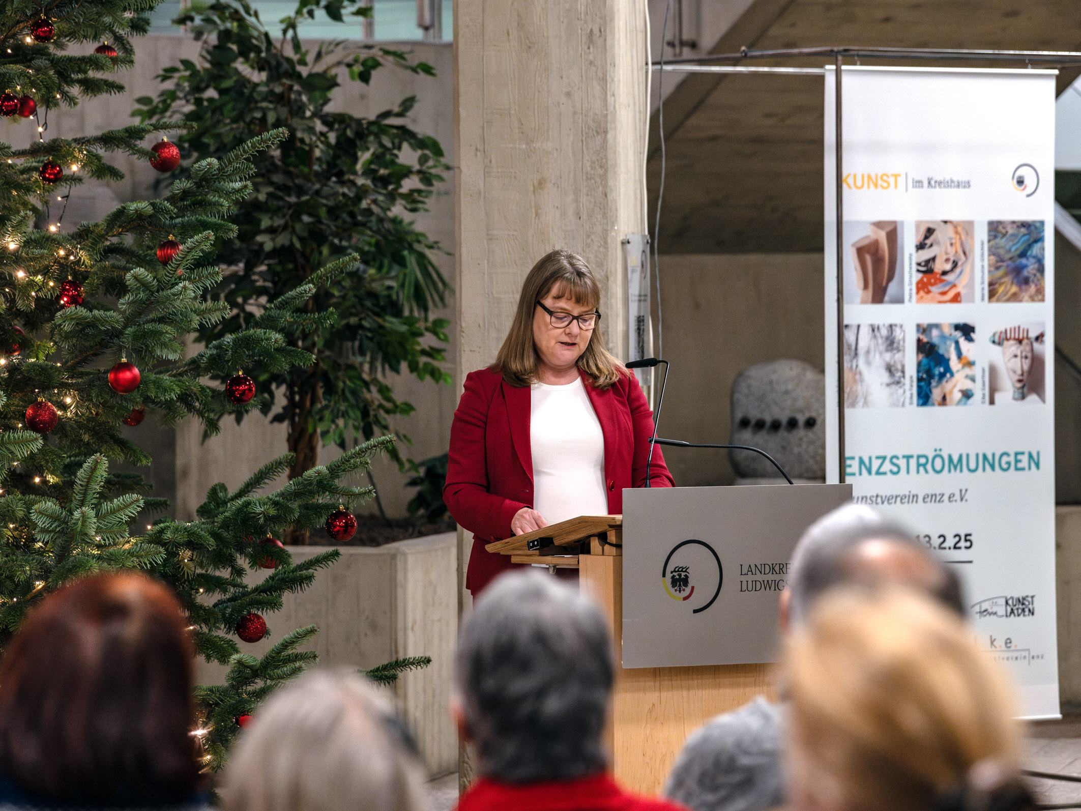 Eine Frau in einem roten Blazer steht am Rednerpult und spricht zu einem Publikum. Im Hintergrund ist ein festlich geschmückter Weihnachtsbaum sichtbar, sowie ein Plakat mit Informationen zur Kunstausstellung „ENZSTRÖMUNGEN“. Der Raum hat eine ruhige, feierliche Atmosphäre.