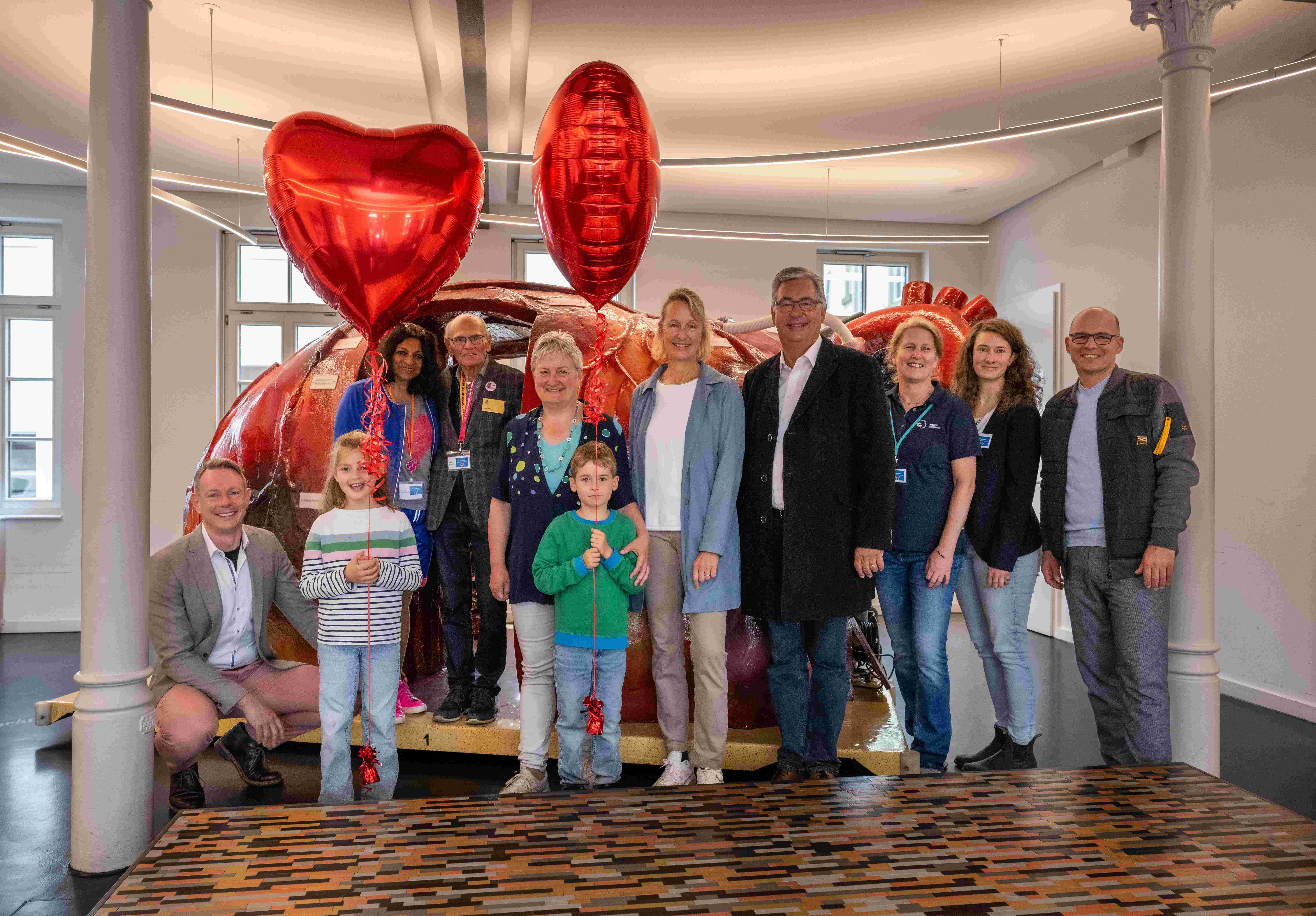 Eine Gruppe von neun Personen steht in einem Raum, umgeben von einer großen, bunten Skulptur. Einige halten Luftballons in Herzform. Die Teilnehmer sind freundlich und lächeln, was eine festliche Atmosphäre vermittelt.