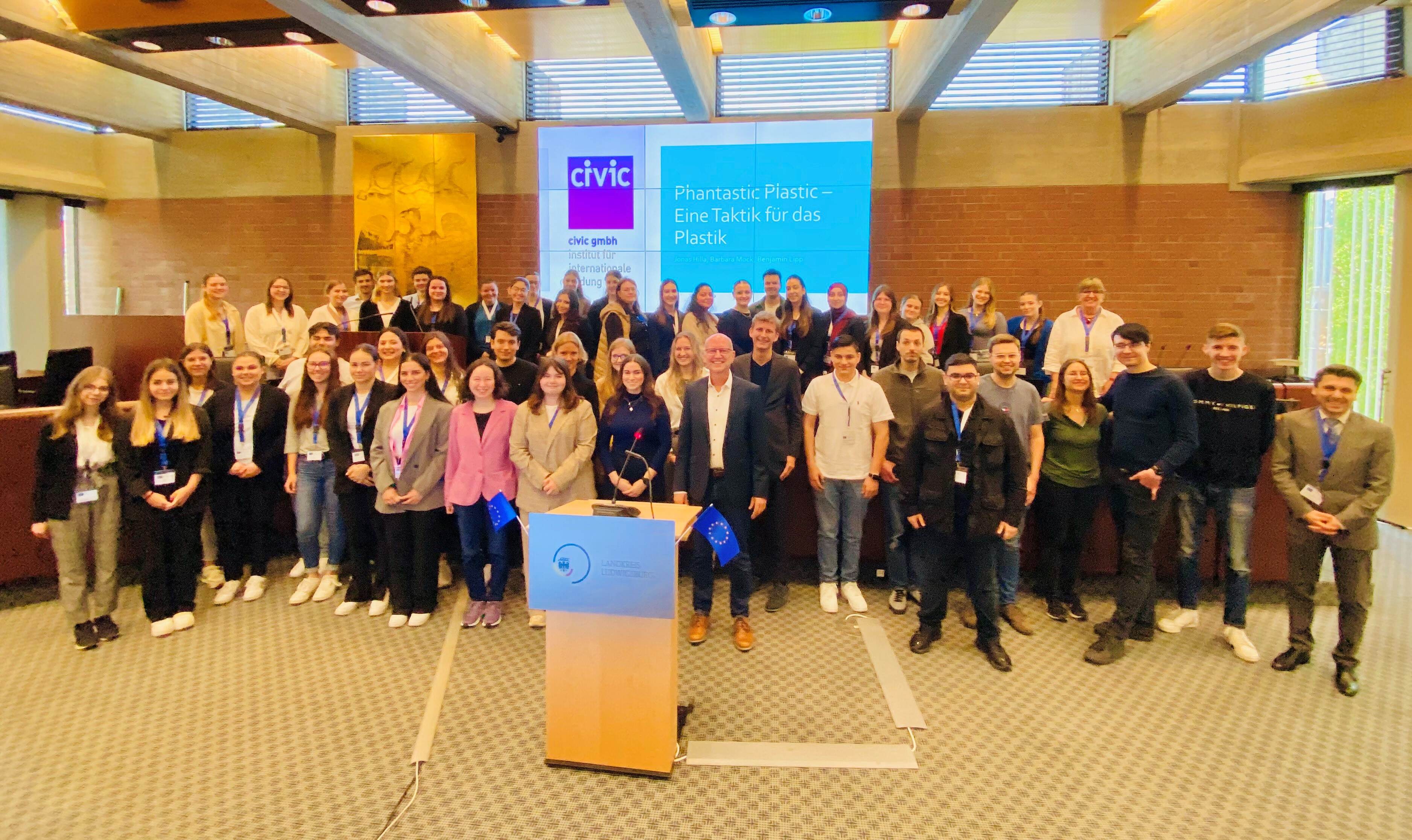 Die Abbildung zeigt eine große Gruppe von Menschen, die in einem Konferenzraum zusammenstehen, um eine Veranstaltung zum Thema Plastik zu besuchen. Im Hintergrund ist ein Bildschirm mit dem Titel "Fantastisch Plastik - Eine Taktik für das Plastik" zu sehen. Das Bild vermittelt eine Atmosphäre des Austauschs und des Engagements.
