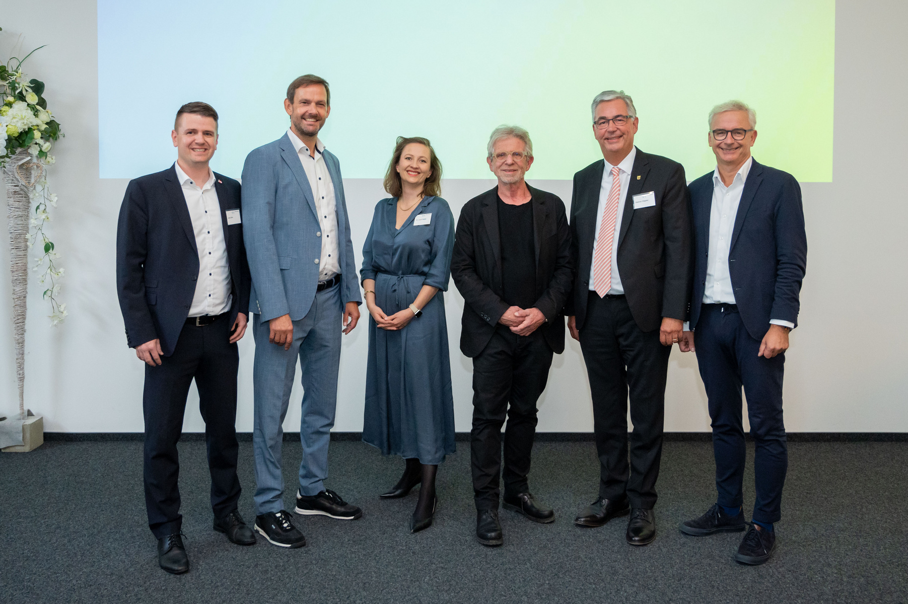 Eine Gruppe von sechs Personen steht lächelnd in einem modernen Raum. Sie tragen formelle Kleidung. Im Hintergrund ist eine hell beleuchtete Wand zu sehen. Das Bild vermittelt eine Atmosphäre der Zusammenarbeit und Professionalität.