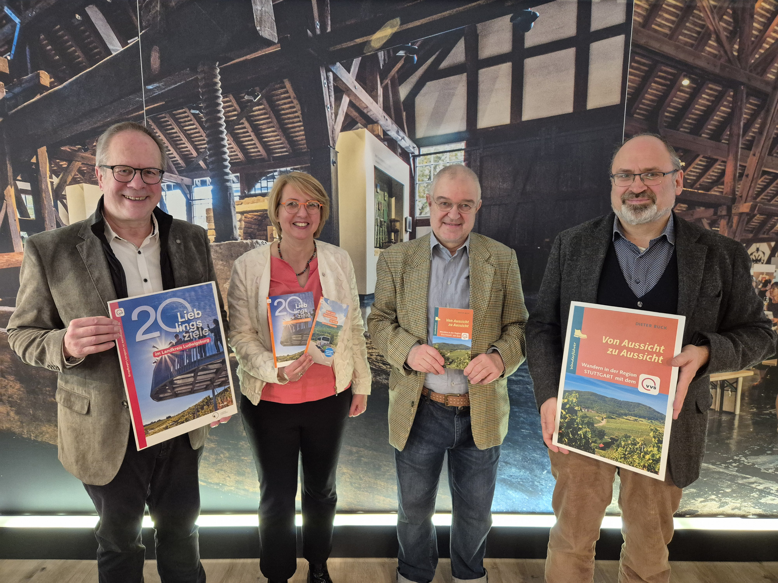 Vier Personen stehen in einem Innenraum, der wie ein historisches Gebäude aussieht. Sie halten verschiedene Broschüren in den Händen, darunter eine mit dem Titel „Von Aussicht zu Aussicht“ und eine andere mit „20 Jahre…“. Die Stimmung ist freundlich und informativ.