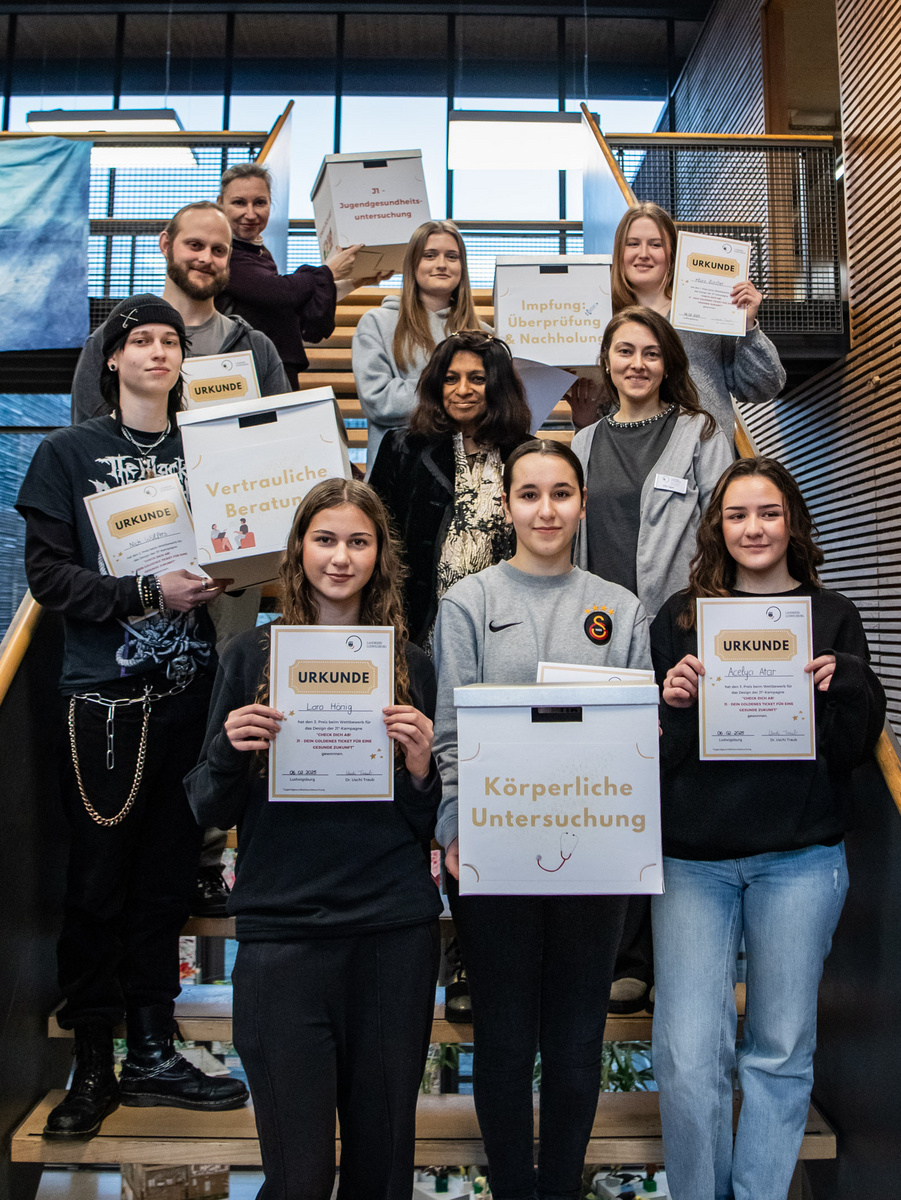 Eine Gruppe von zehn Personen steht auf einer Treppe und hält Zertifikate in den Händen. Die Themen der Zertifikate sind vertrauliche Beratung und körperliche Untersuchung. Im Hintergrund sind Holzgestelle und Fenster zu sehen. Die Stimmung ist feierlich und stolz.