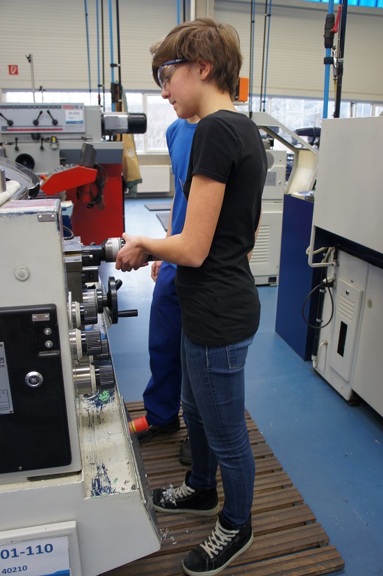 Eine junge Frau steht an einer Maschine in einer Werkstatt und bedient diese. Sie trägt ein schwarzes T-Shirt und eine Schutzbrille. Im Hintergrund ist eine andere Person zu sehen, die ebenfalls an der Maschine arbeitet.