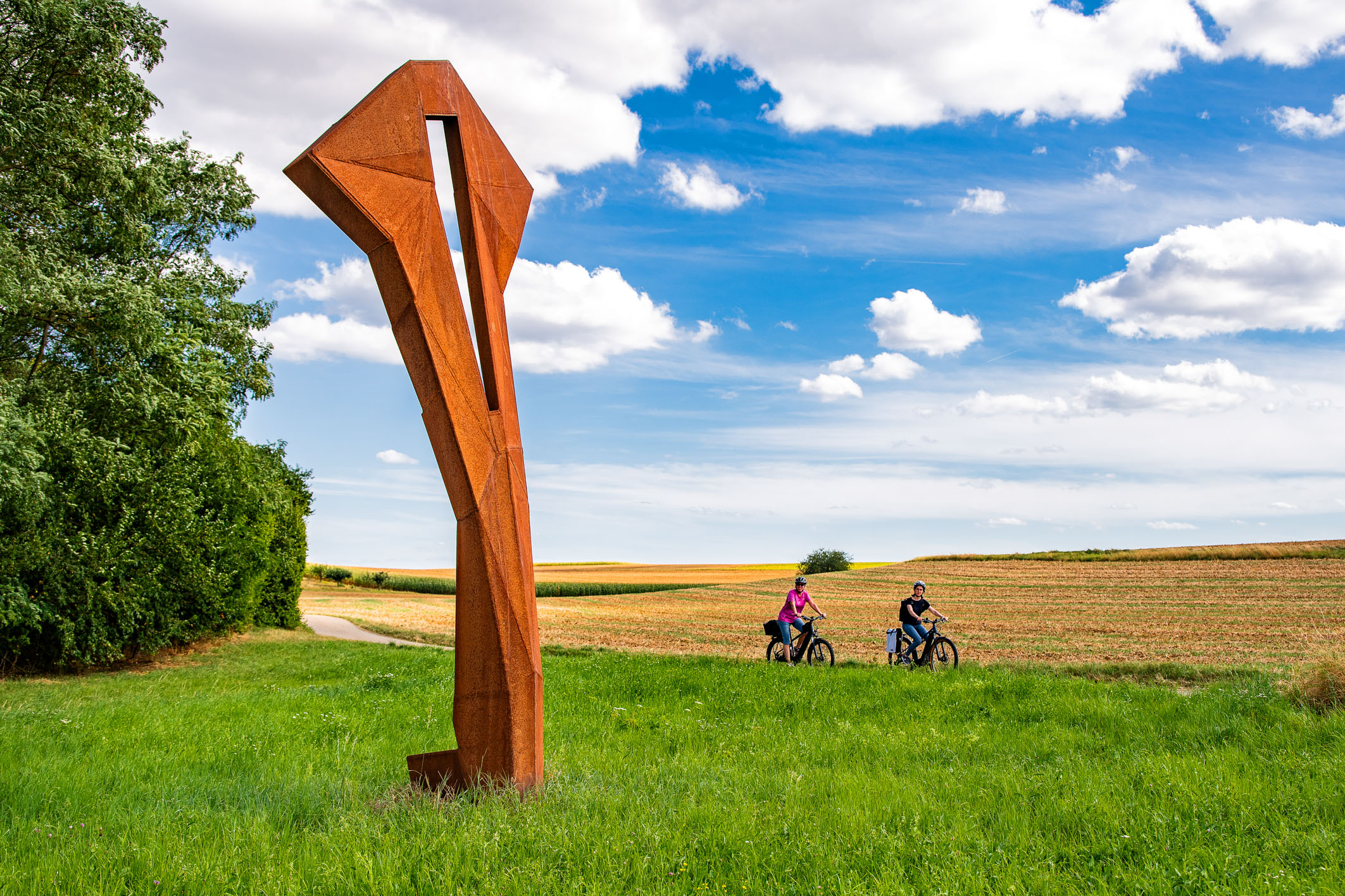A tall, abstract, rust-colored sculpture stands in a grassy area. Two cyclists pass by on a path, surrounded by fields and a blue sky with clouds, creating a serene outdoor atmosphere.