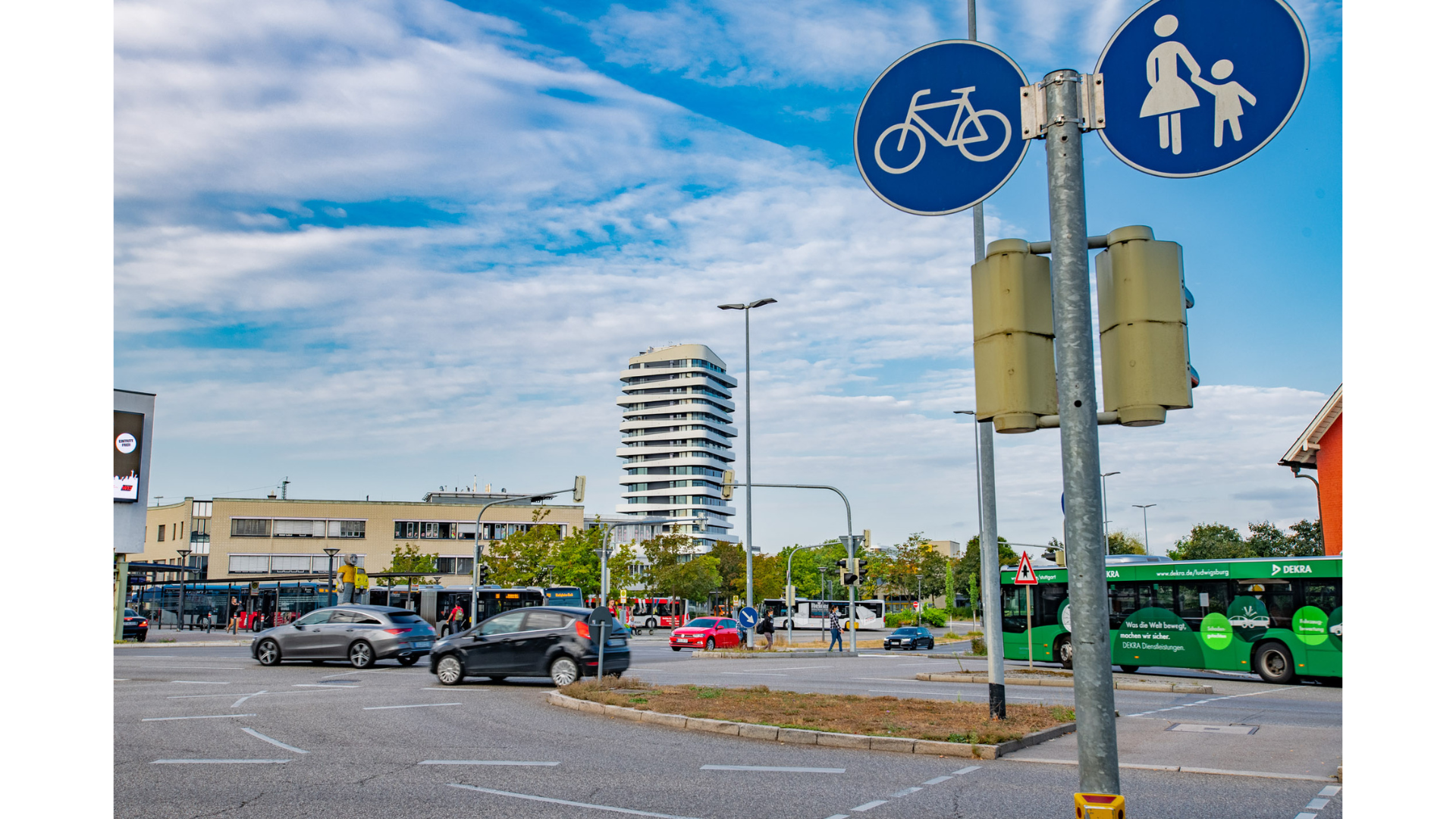Das Bild zeigt eine Verkehrsinszenierung an einem Kreisverkehr mit Verkehrsschildern für Radfahrer und Fußgänger. Im Hintergrund sind Busse, Autos und ein modernes Gebäude zu sehen, unter blauem Himmel mit teilweise Wolken.