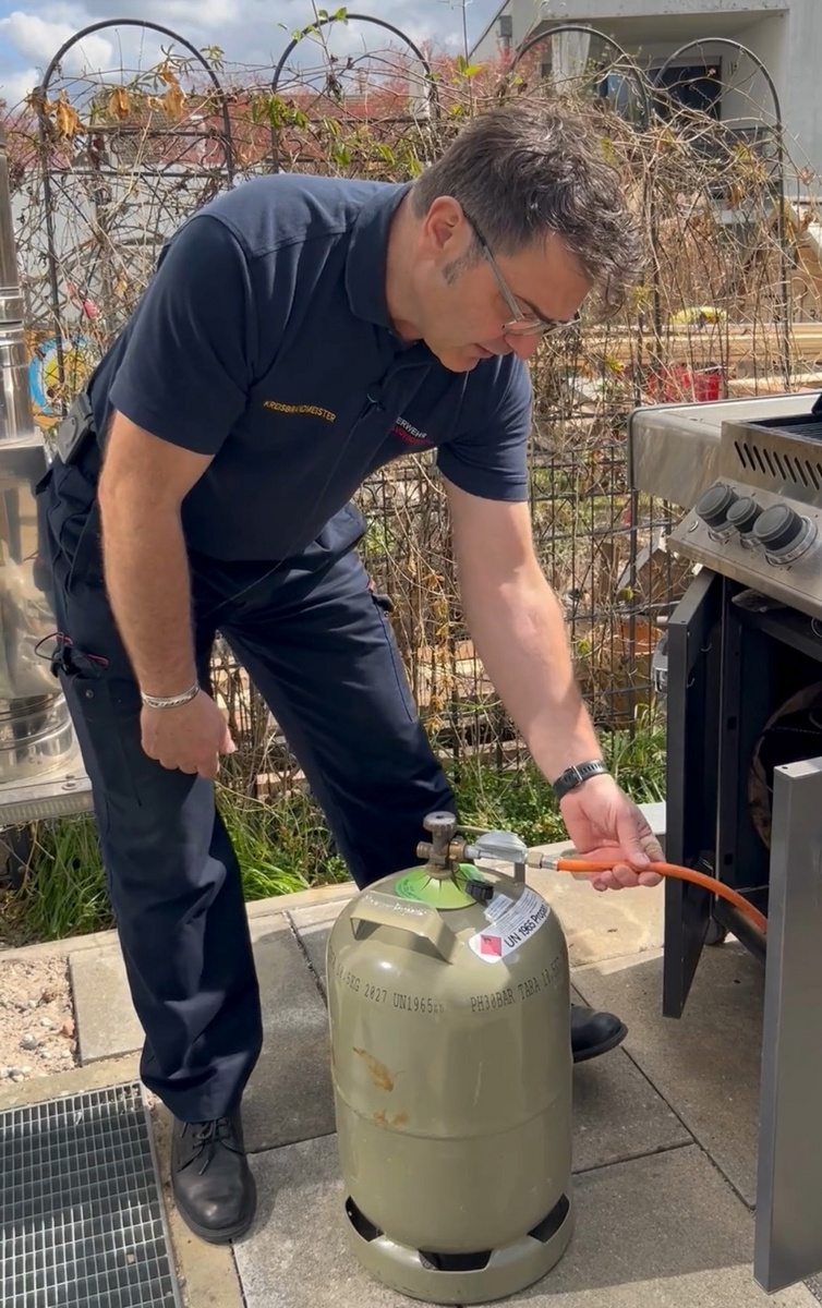 Eine Person in Arbeitskleidung beugt sich über einen Gaszylinder, während sie einen Schlauch daran anschließt. Der Hintergrund zeigt einen Grill und eine Freizeitumgebung im Freien.