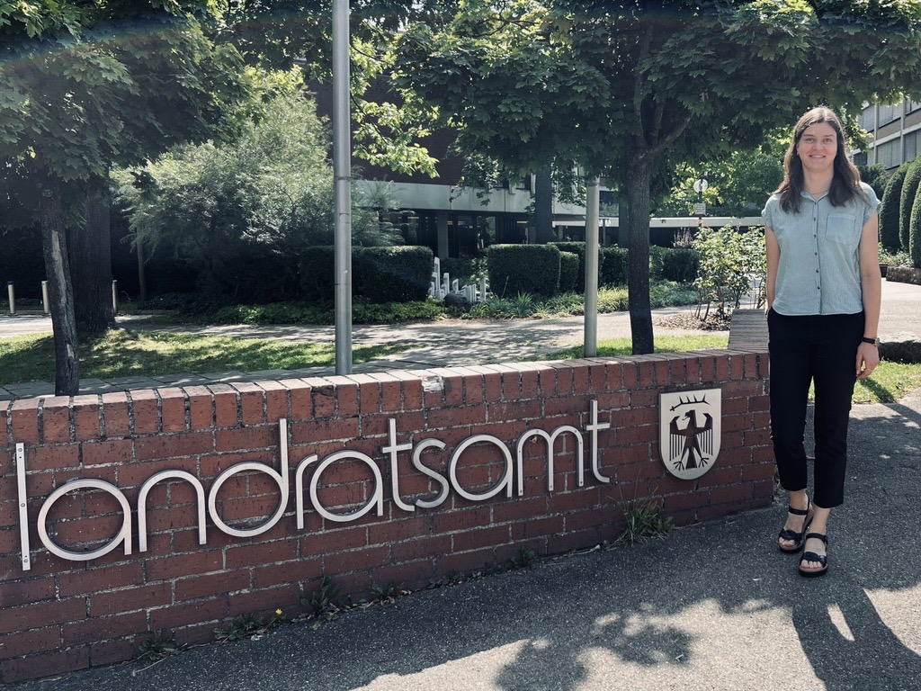 Eine Person steht neben dem Schild des Landratsamts. Das Schild zeigt den Schriftzug "landratsamt" und ein Wappen. Im Hintergrund sind Bäume und ein Gebäude sichtbar. Es ist ein sonniger Tag.