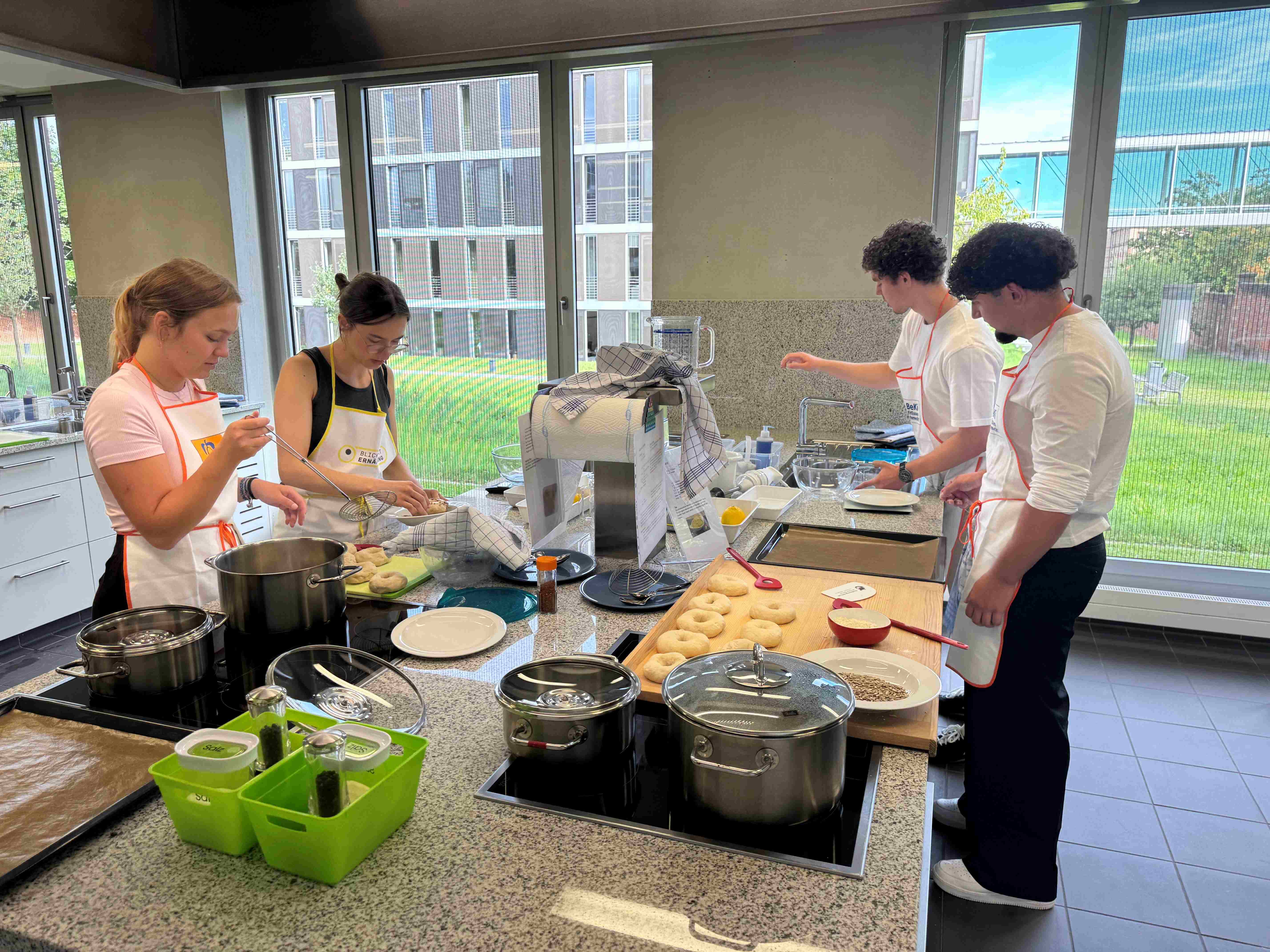 Vier Personen arbeiten in einer modernen Küche. Sie bereiten verschiedene Lebensmittel vor und verwenden Kochutensilien. Auf der Arbeitsfläche sind Töpfe, Schüsseln und Zutaten sichtbar. Ein Fenster im Hintergrund zeigt den Außenbereich.