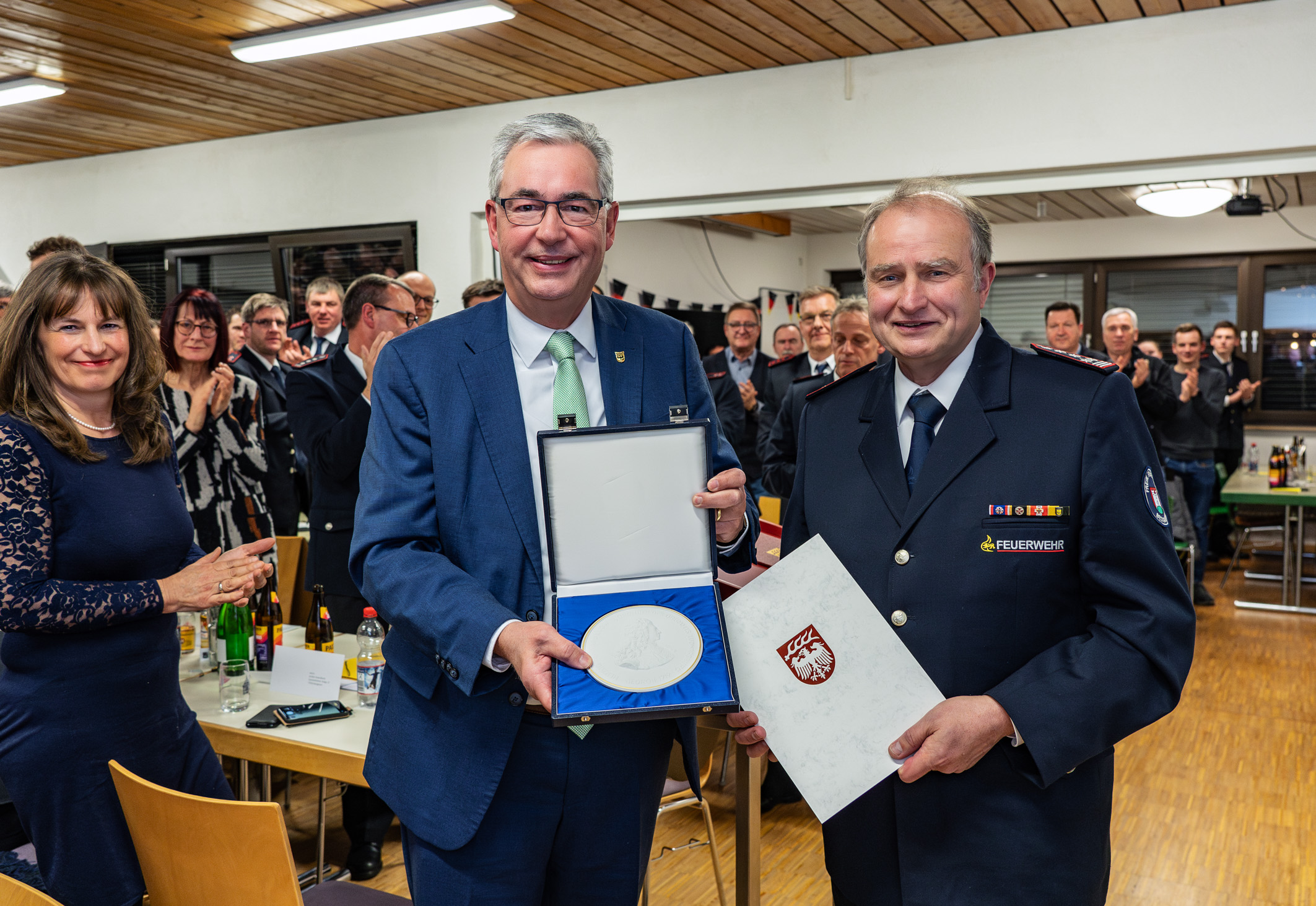 Zwei Männer stehen im Vordergrund und halten eine Auszeichnung in einer Schatulle. Der Mann links trägt einen Anzug und Brille, während der Mann rechts eine Feuerwehruniform trägt. Im Hintergrund ist eine Gruppe von applaudierenden Menschen zu sehen, die eine feierliche Stimmung vermittelt.