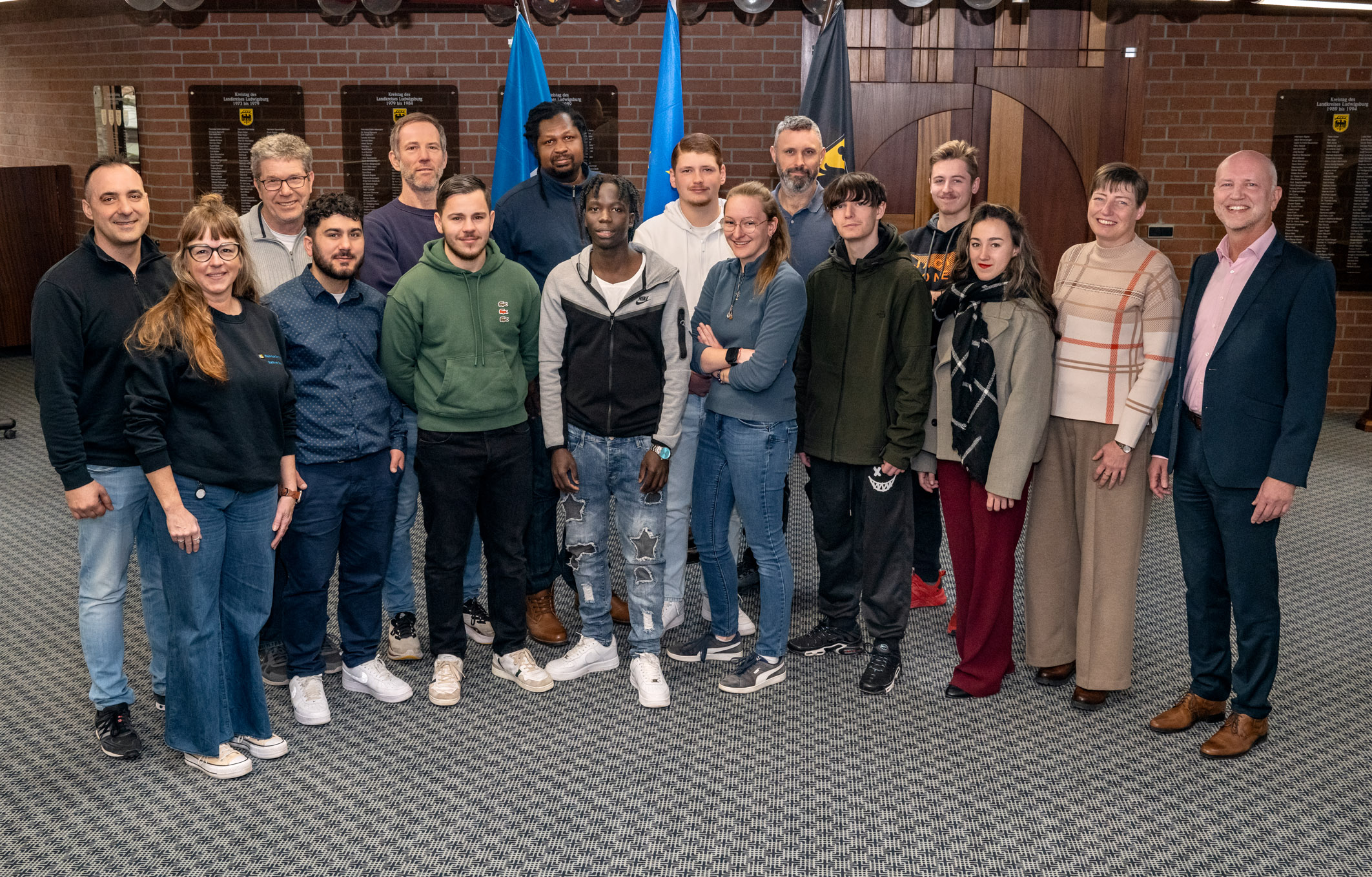 Eine Gruppe von 16 Personen steht in einem Raum zusammen, unterstützt von verschiedenen Flaggen im Hintergrund. Die Anwesenden sind unterschiedlich gekleidet und lächeln in die Kamera. Die Atmosphäre wirkt freundlich und informell, möglicherweise handelt es sich um ein Treffen oder eine Veranstaltung.