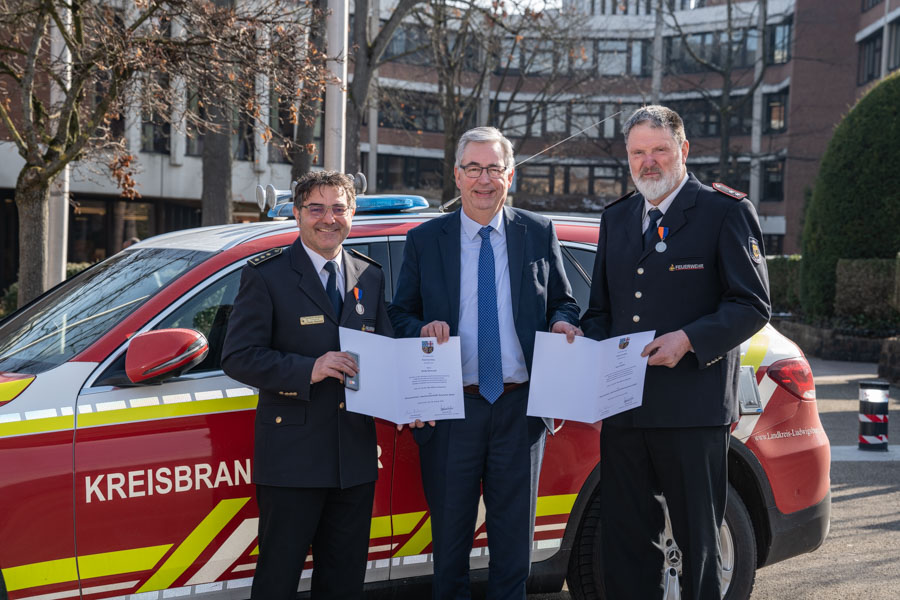 Drei Männer stehen vor einem Einsatzfahrzeug der Feuerwehr. Zwei von ihnen halten Urkunden in den Händen, während der dritte in der Mitte steht und lächelt. Alle tragen formelle Kleidung, und im Hintergrund sind Bäume und Gebäude zu sehen.