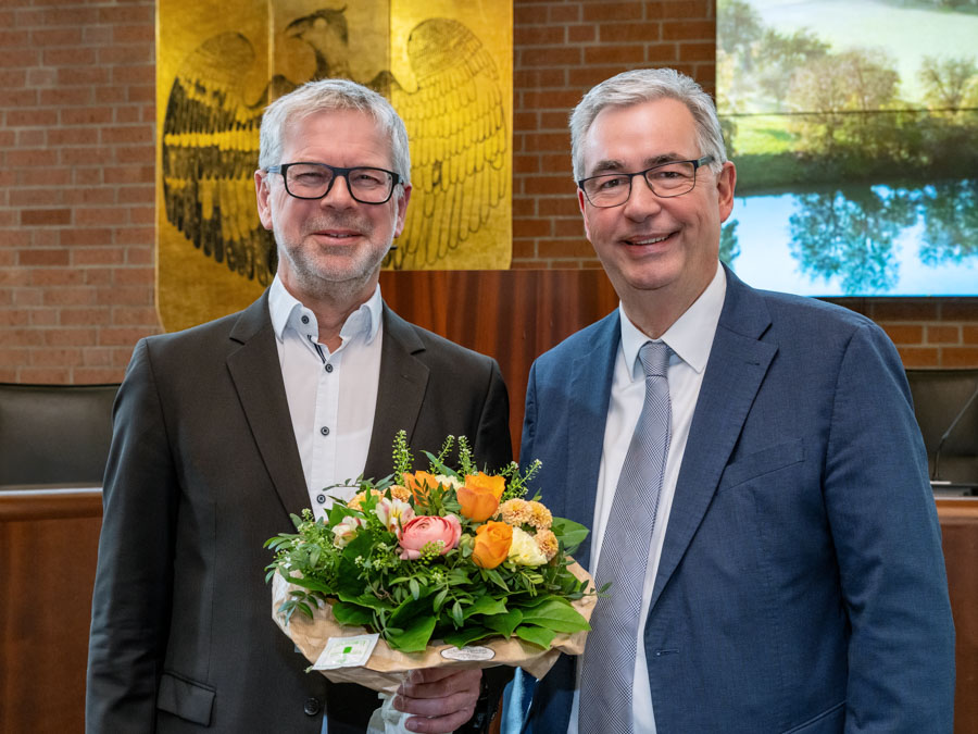 Zwei Männer stehen lächelnd in einem offiziellen Raum. Der Mann links trägt eine Brille und hält einen bunten Blumenstrauß. Der Mann rechts trägt einen blauen Anzug. Im Hintergrund ist eine Wand mit einem Wappen und einem Bild von Landschaften sichtbar.