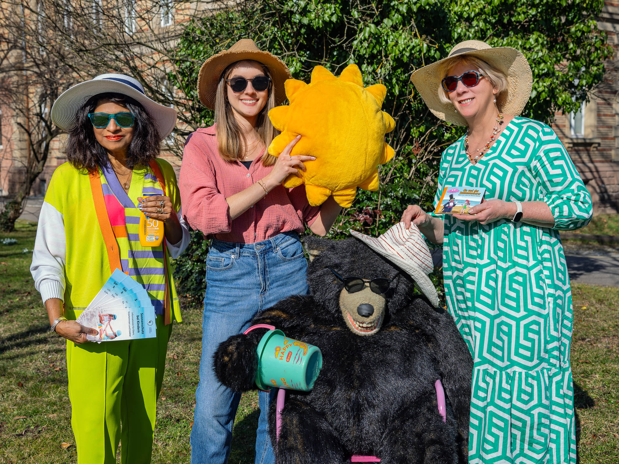 Drei Frauen stehen im Freien und lächeln. Eine Frau hält eine gelbe Sonne, während die andere ein Plüschtierbär mit Hut präsentiert. Die Frauen tragen Sonnenhüte und gestalten einen fröhlichen, sommerlichen Eindruck. Sie haben Flyer oder Broschüren in der Hand.