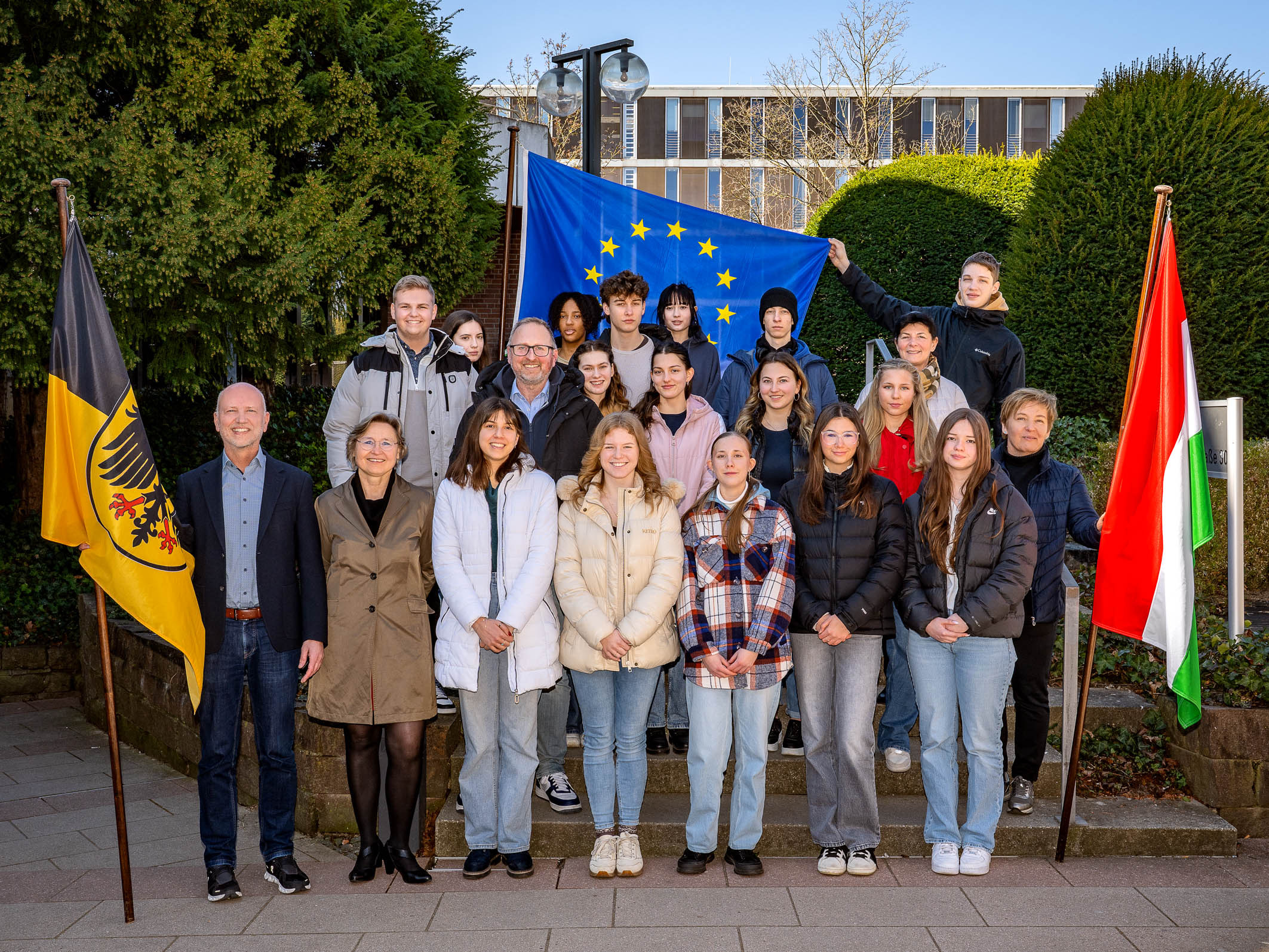 Eine Gruppe von etwa 20 Personen steht draußen vor grünen Hecken. Im Hintergrund ist eine europäische Flagge zu sehen. Links und rechts der Gruppe stehen Fahnen. Die Anwesenden lächeln und scheinen an einem gemeinsamen Projekt teilzunehmen.