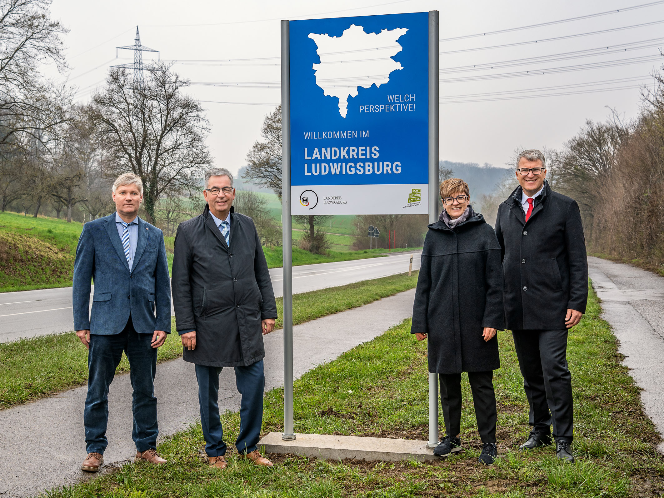 Die Aufnahme zeigt vier Personen neben einem Willkommensschild, das "Willkommen im Landkreis Ludwigsburg" anzeigt. Im Hintergrund sind eine Straße und Bäume zu sehen. Die Personen tragen formelle Kleidung und stehen auf einer Wiese.