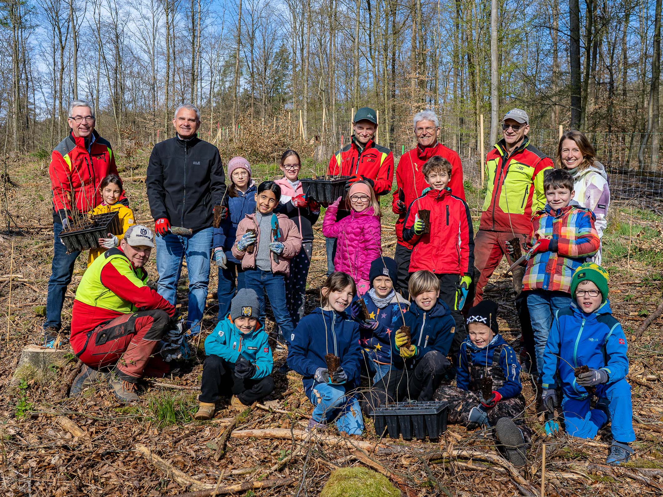Eine Gruppe von Kindern und Erwachsenen steht im Wald und hält Setzlinge in der Hand. Sie lächeln und posieren gemeinsam für ein Foto, während einige in Arbeitshosen und -jacken gekleidet sind. Die Szene vermittelt ein Gefühl von Gemeinschaft und Engagement für das Pflanzen von Bäumen.