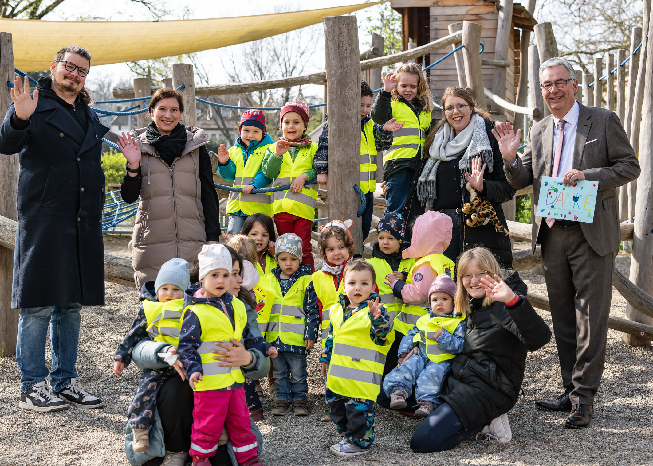 Eine Gruppe von Kindern in leuchtenden Sicherheitswesten steht auf einem Spielplatz. Sie sind umgeben von Erwachsenen, die lächeln und die Hände zum Winken heben. Die Szene vermittelt Freude und Gemeinschaft. Im Hintergrund sind Spielgeräte sichtbar.