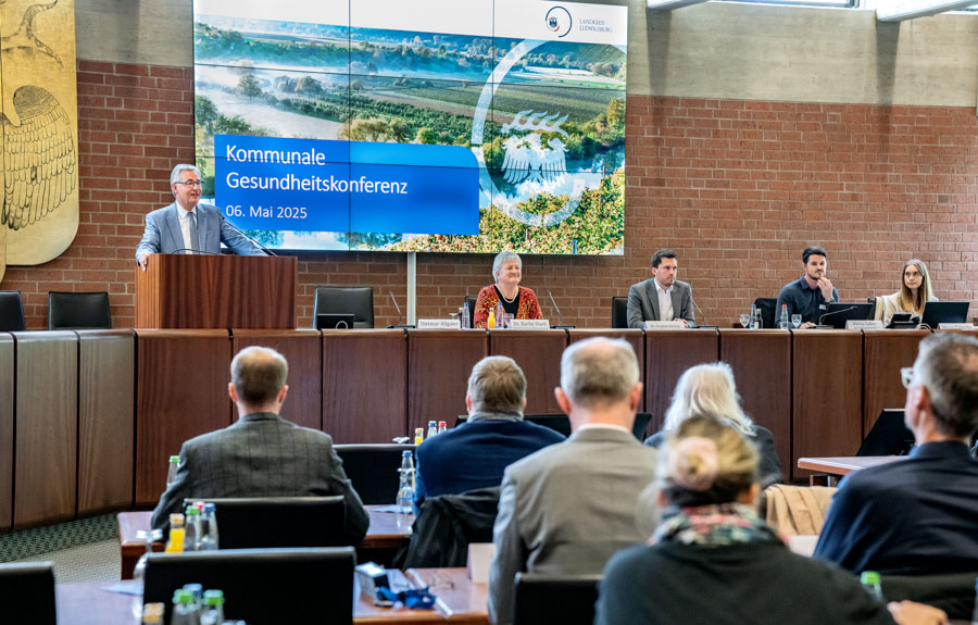 Bildbeschreibung: Eine Person steht am Rednerpult auf einer Konferenz zum Thema Gesundheit. Auf einem großen Bildschirm im Hintergrund steht „Kommunale Gesundheitskonferenz, 06. Mai 2025“. Die Zuschauer im Raum hören aufmerksam zu.