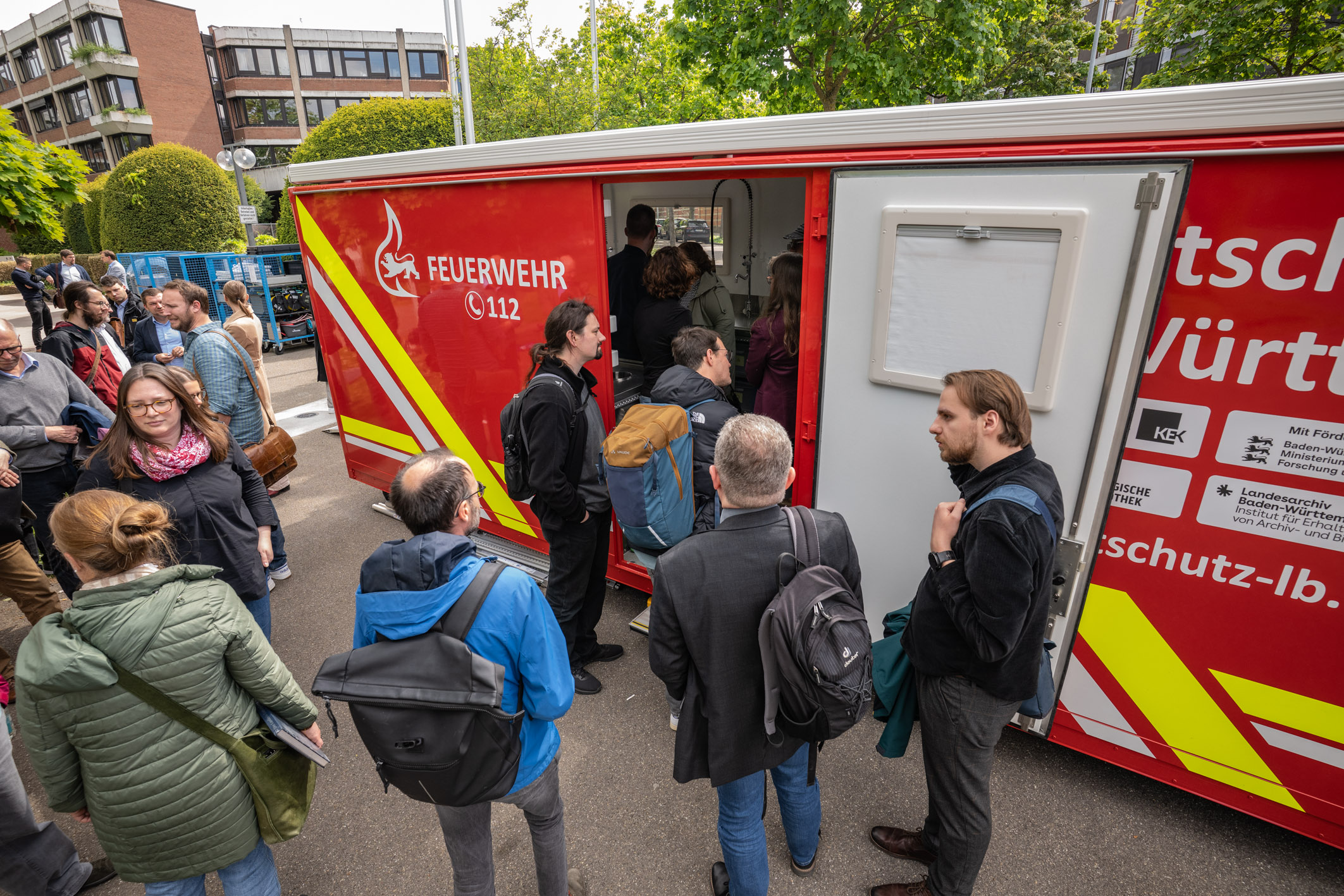 Eine Gruppe von Menschen steht vor einem Feuerwehrfahrzeug. Einige Personen sind im Gespräch, während andere in Richtung des Fahrzeugs schauen. Das Fahrzeug hat eine rote Außenseite mit dem Schriftzug "FEUERWEHR" und der Notrufnummer 112.