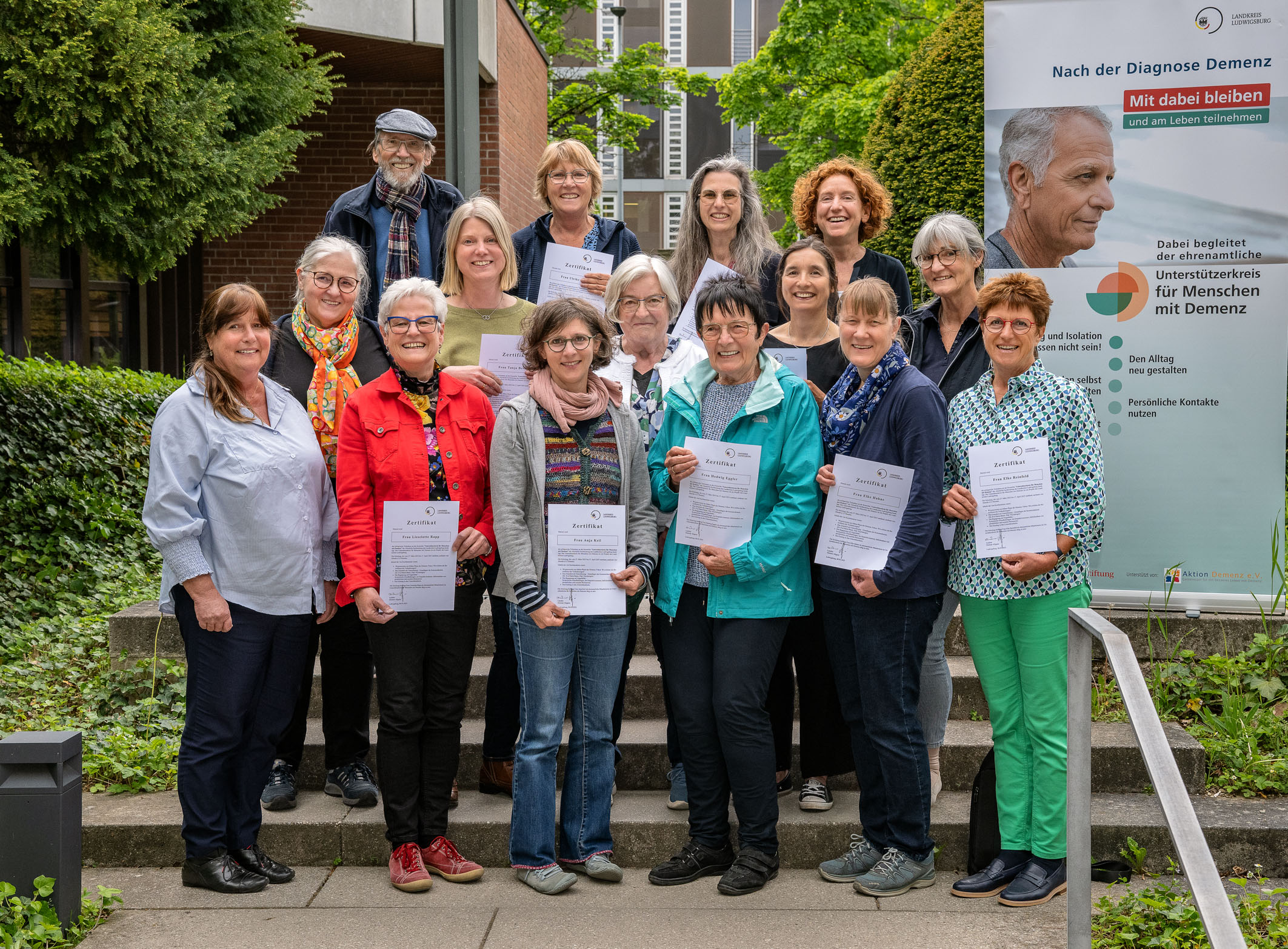 Eine Gruppe von 16 Personen steht auf einer Treppe im Freien, lächelt und hält Dokumente in den Händen. Im Hintergrund ist ein Banner mit dem Text „Nach der Diagnose Demenz - Mit dabei bleiben“ zu sehen. Das Bild vermittelt ein Gefühl von Unterstützung und Gemeinschaft.