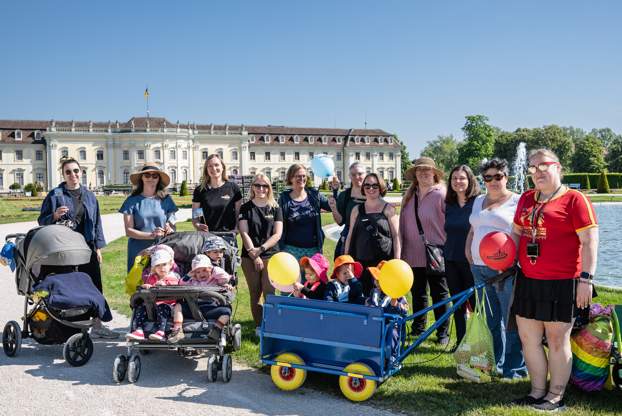 Eine Gruppe von Frauen und Kindern steht in einem Park vor einem beeindruckenden Gebäude. Einige Frauen tragen Sonnenhüte, und im Vordergrund sind Kinder in Kinderwagen und ein Wagen mit Luftballons zu sehen. Der Himmel ist klar und blau.