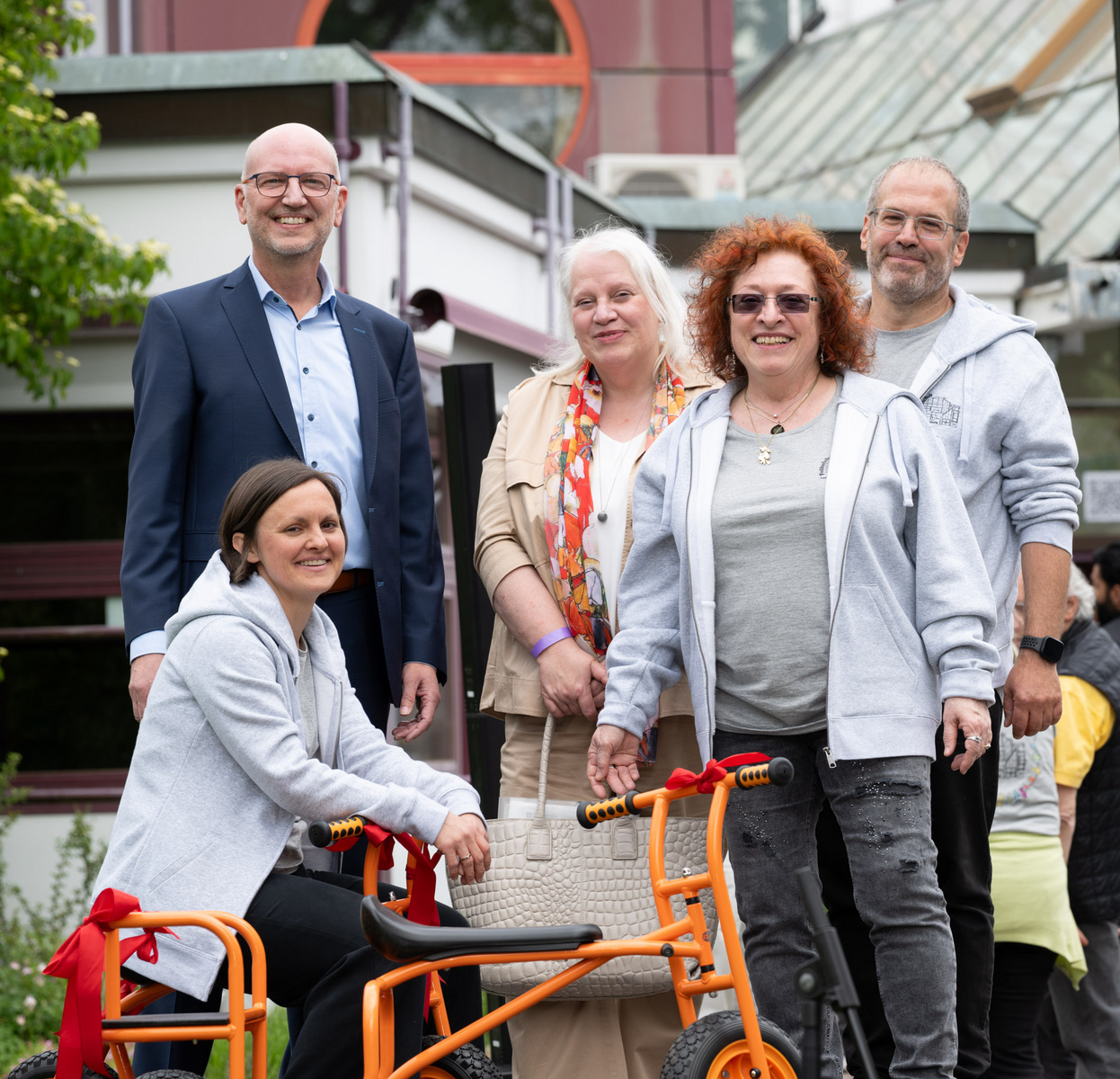 Sechs Personen stehen zusammen im Freien. Eine Frau sitzt auf einem orangefarbenen Kinderfahrrad. Sie lächeln und wirken glücklich. Es gibt eine grüne Umgebung im Hintergrund, einschließlich Bäume und Gebäude.