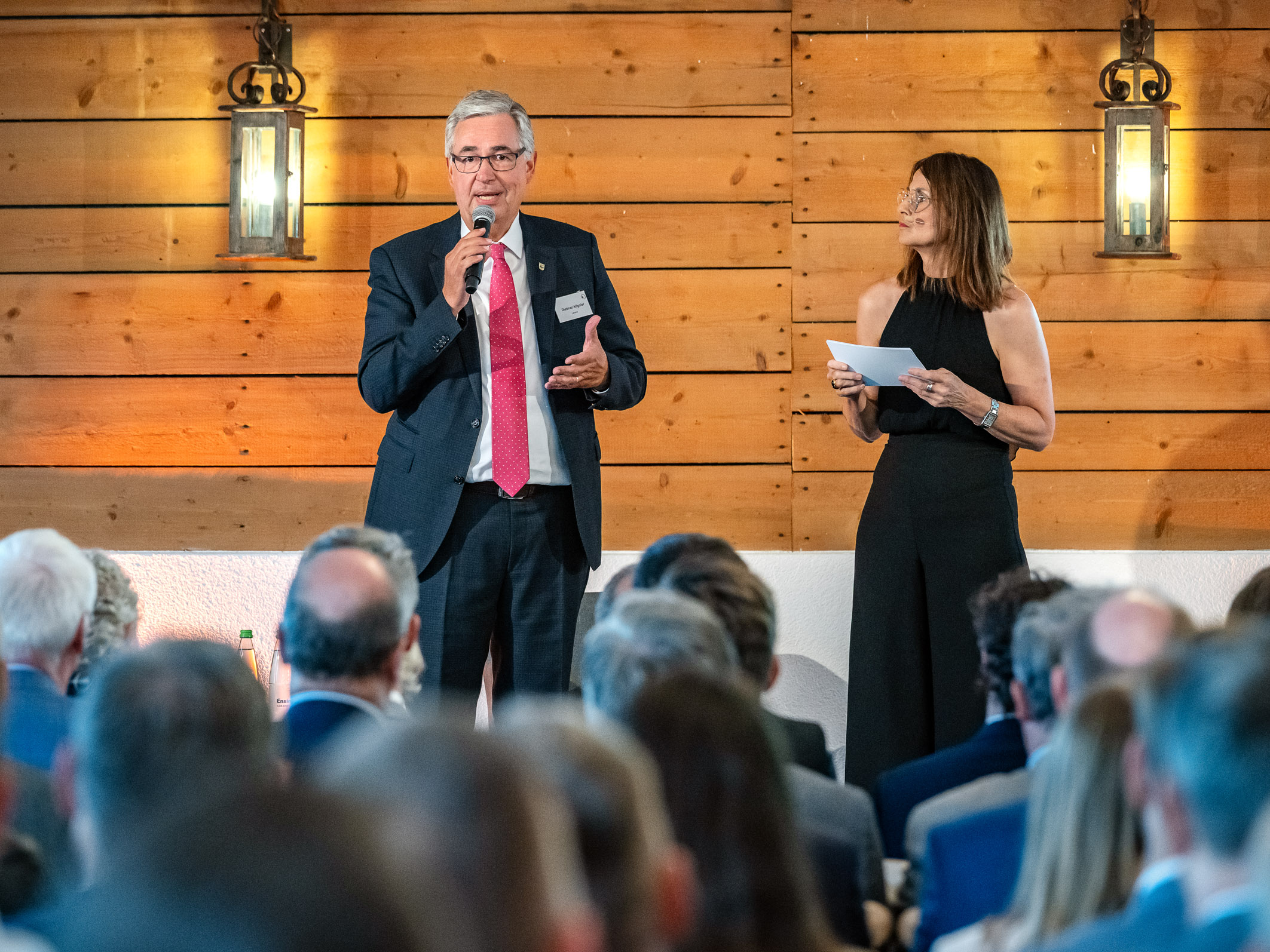 Ein Mann spricht mit einem Mikrofon in der Hand, während eine Frau neben ihm steht und Notizen in der Hand hält. Sie befinden sich auf einer Bühne vor einem Publikum in einem festlichen Raum mit Holzwänden und beleuchtetem Ambiente.