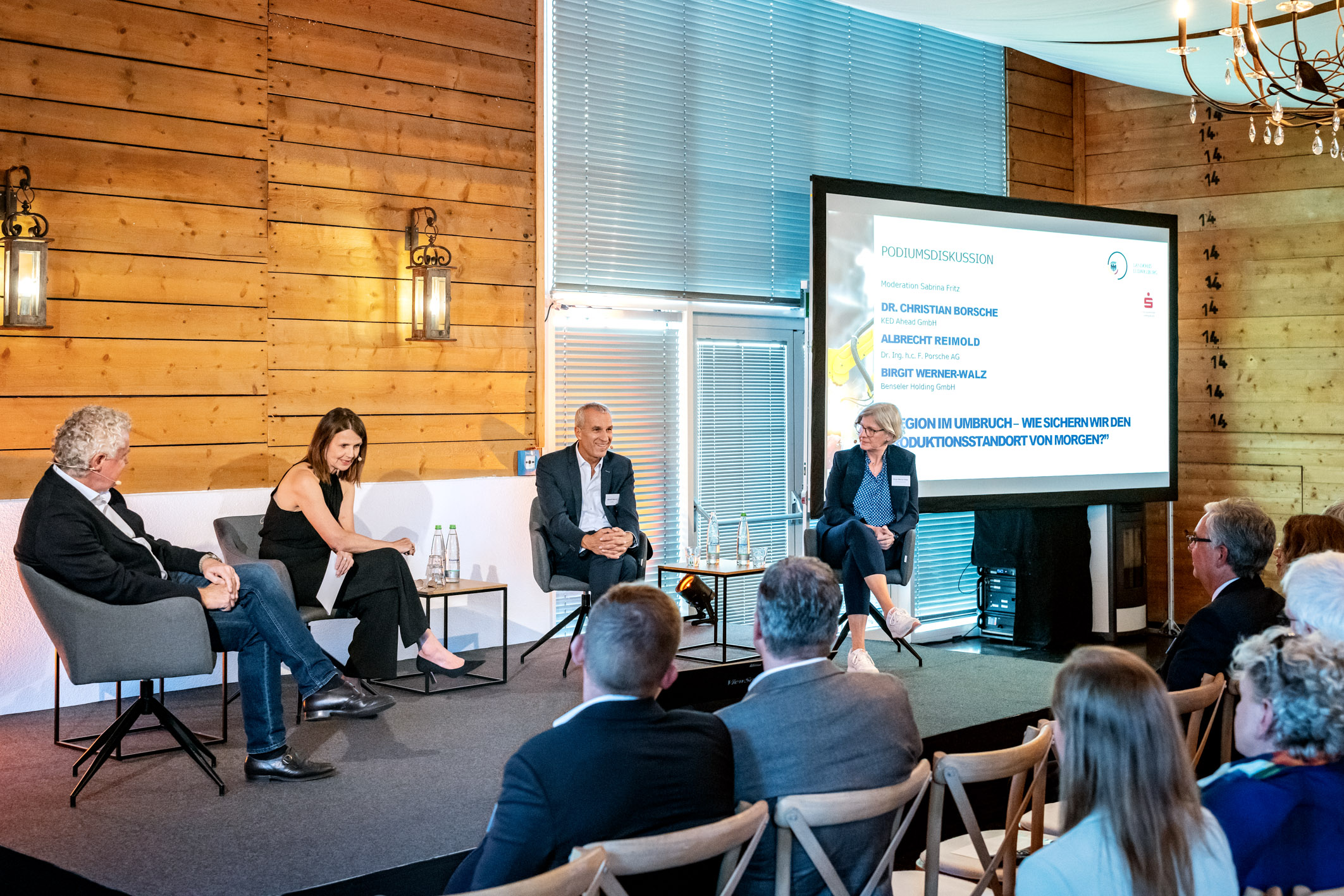 Eine Podiumsdiskussion mit drei Sprechern auf einem Szenenpodium. Im Hintergrund ist eine Präsentation sichtbar. Die Umgebung hat eine rustikale Holzgestaltung und es sitzen Zuschauer in Stühlen, die auf das Podium schauen.