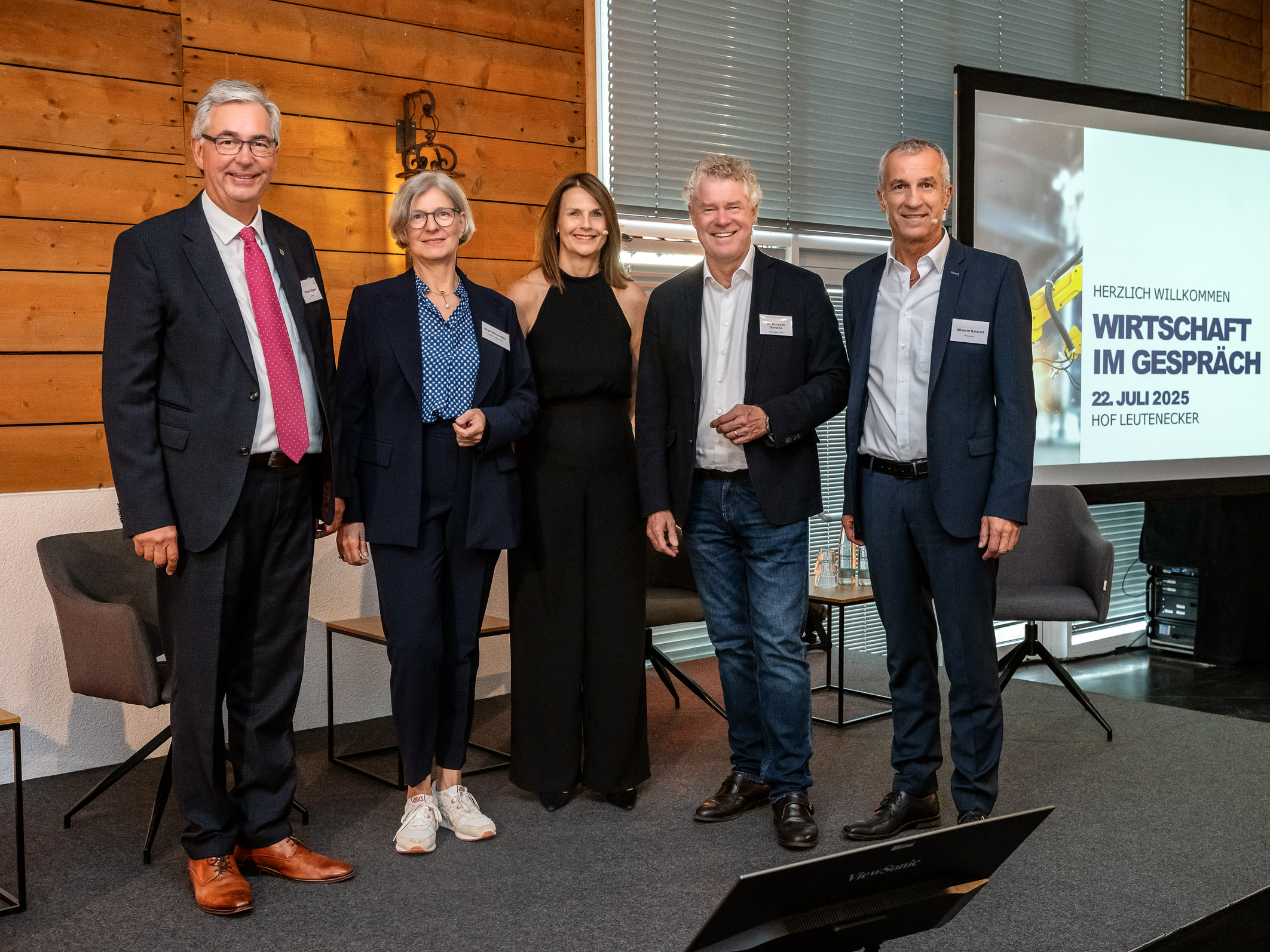 Fünf Personen stehen nebeneinander in einem Raum. Sie lächeln und tragen formelle Kleidung. Im Hintergrund ist ein Bildschirm sichtbar, der eine Einladung zu einer Veranstaltung mit dem Titel "Wirtschaft im Gespräch" anzeigt, datiert auf den 22. Juli 2023.