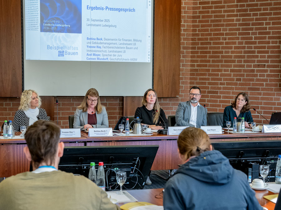 A panel of five speakers sits at a conference table during a press event. They appear to be discussing subjects related to education and finance. A large screen displays information above them. Attendees are seated in the foreground, focusing on the speakers.