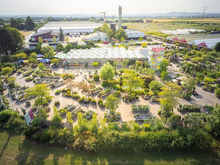 Die Luftaufnahme zeigt einen Garten mit vielen Pflanzen, Bäumen und Blumenbeeten. Im Hintergrund sind Gewächshäuser und Lagerhallen zu sehen. Der Garten ist ordentlich angelegt und lädt zum Spaziergang ein. Die Umgebung wirkt ruhig und grün, ideal für Gartenliebhaber.