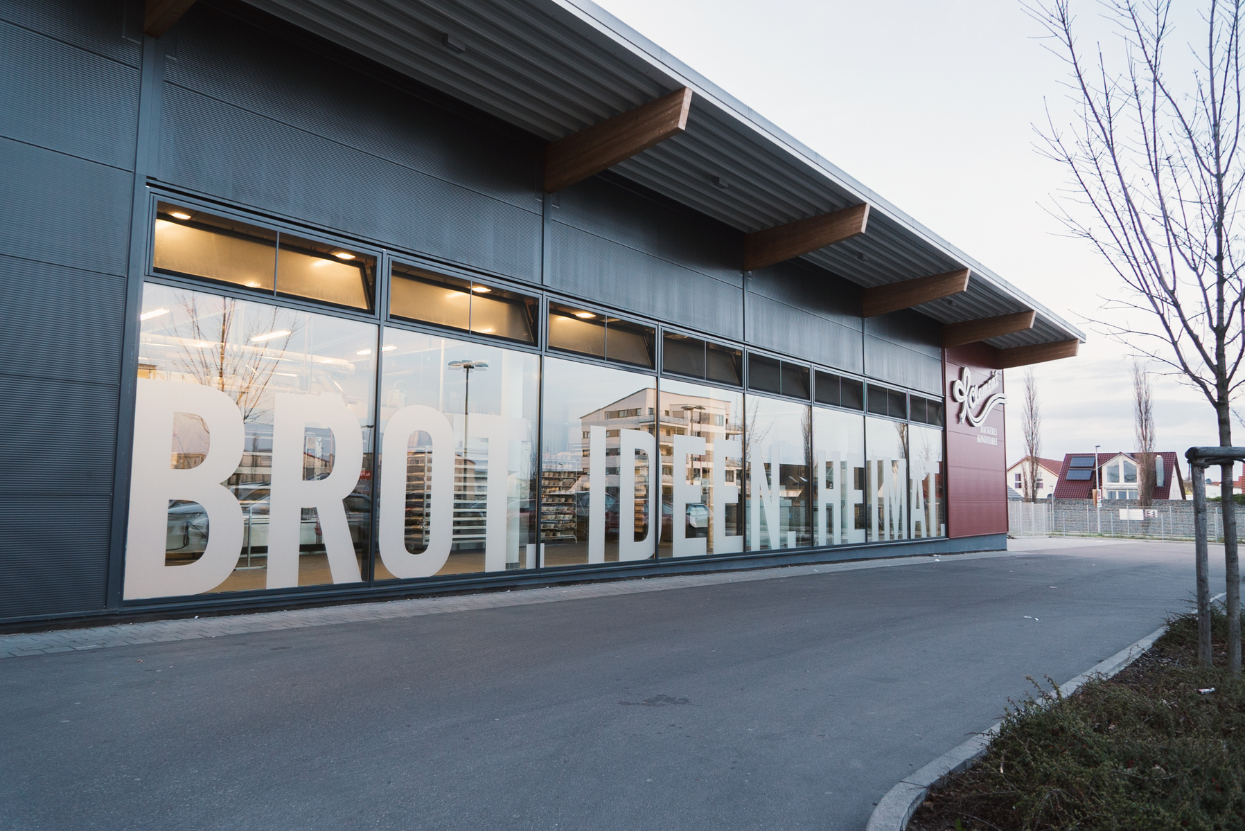 Die Außenansicht eines modernen Gebäudes mit großen Fenstern, auf denen die Worte "BROTHEIDEEN HEIMAT" in großen Buchstaben zu lesen sind. Vor dem Gebäude ist ein gepflasterter Weg und einige Sträucher im Vordergrund.