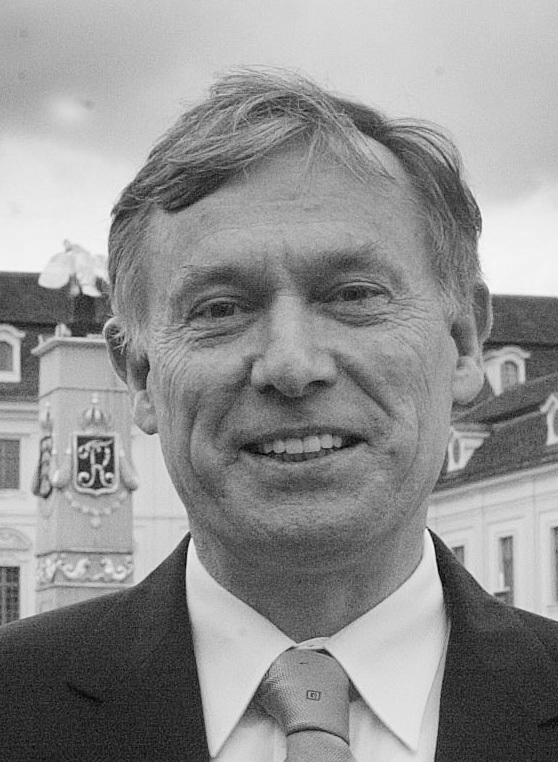 Ein Mann mit kurzen, grauen Haaren lächelt freundlich in die Kamera. Er trägt einen dunklen Anzug und eine helle Krawatte. Im Hintergrund sind unscharfe historische Gebäude zu erkennen, die zur Atmosphäre des Ortes beitragen.