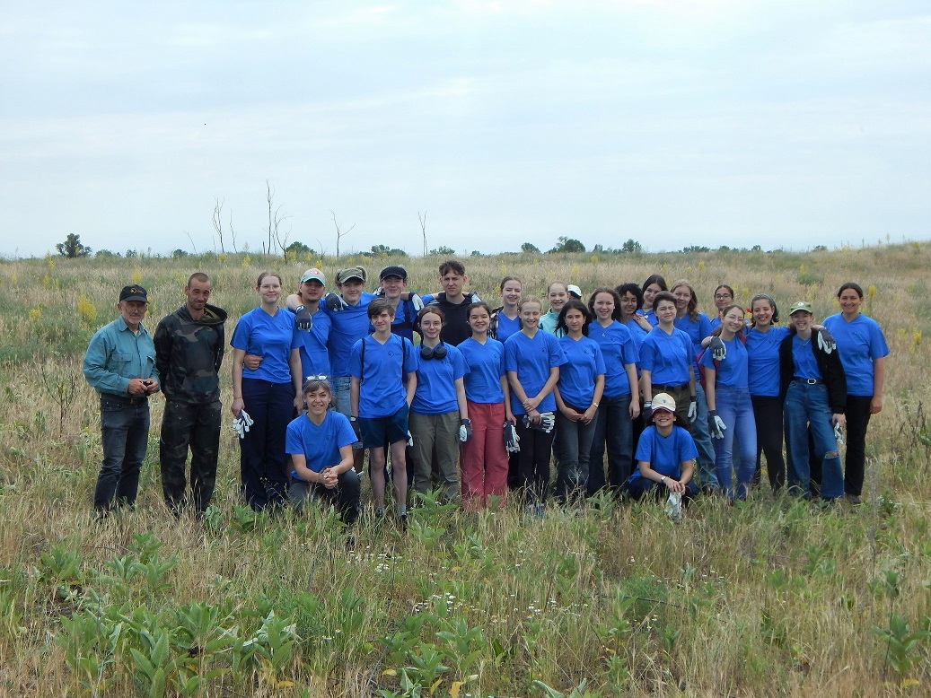 Eine Gruppe von etwa 30 Menschen steht lächelnd auf einer offenen Wiese. Die meisten tragen blaue T-Shirts. Die Umgebung ist ländlich, mit etwas hohem Gras und wenigen Bäumen im Hintergrund. Der Himmel ist bewölkt.