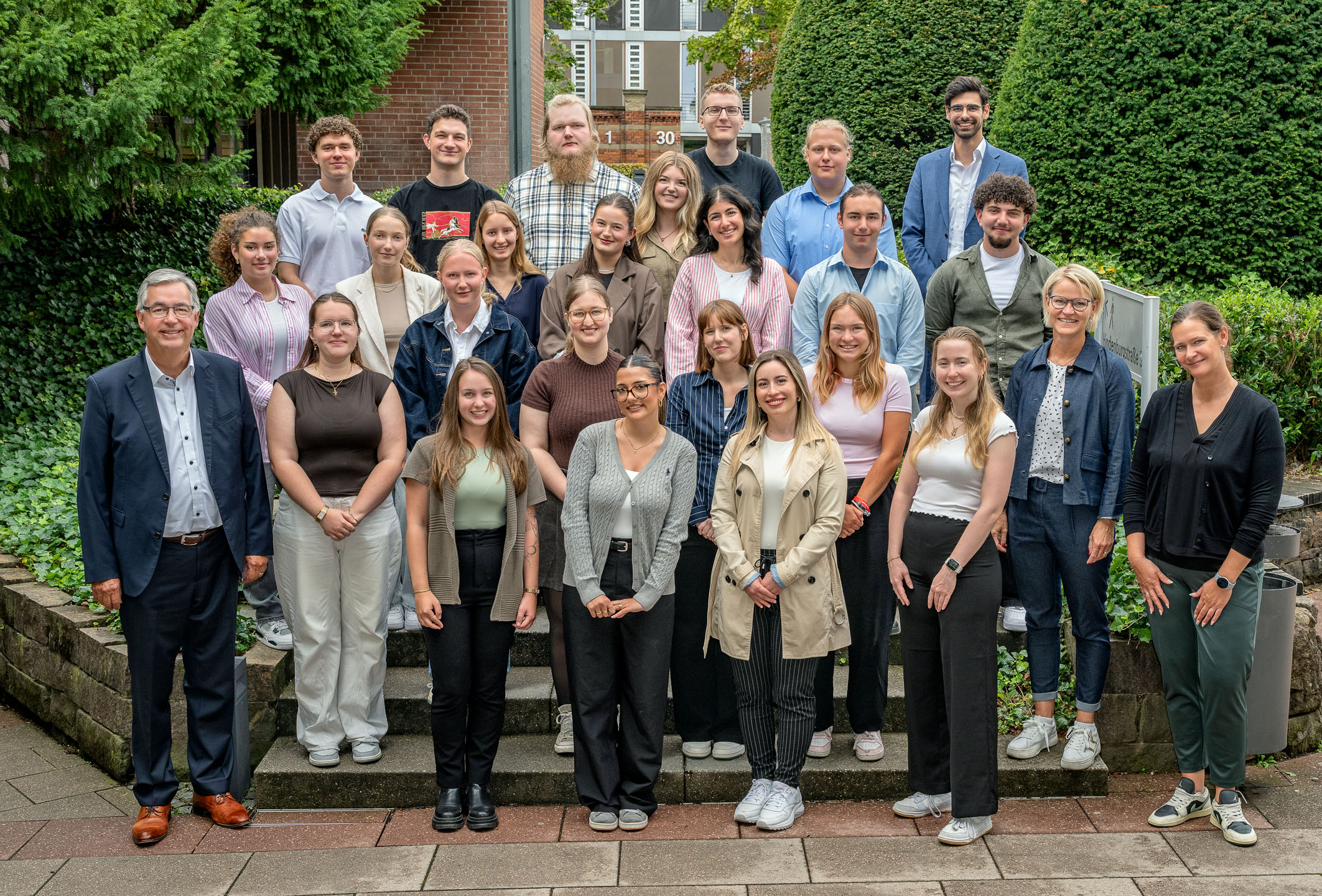 Eine Gruppe von etwa 30 Personen steht auf einer Treppe in einem Garten. Sie sind in professioneller Kleidung. Im Vordergrund steht ein älterer Mann mit Brille und zwei Frauen in hellem und dunklem Outfit. Die Atmosphäre ist freundlich und gemeinschaftlich.