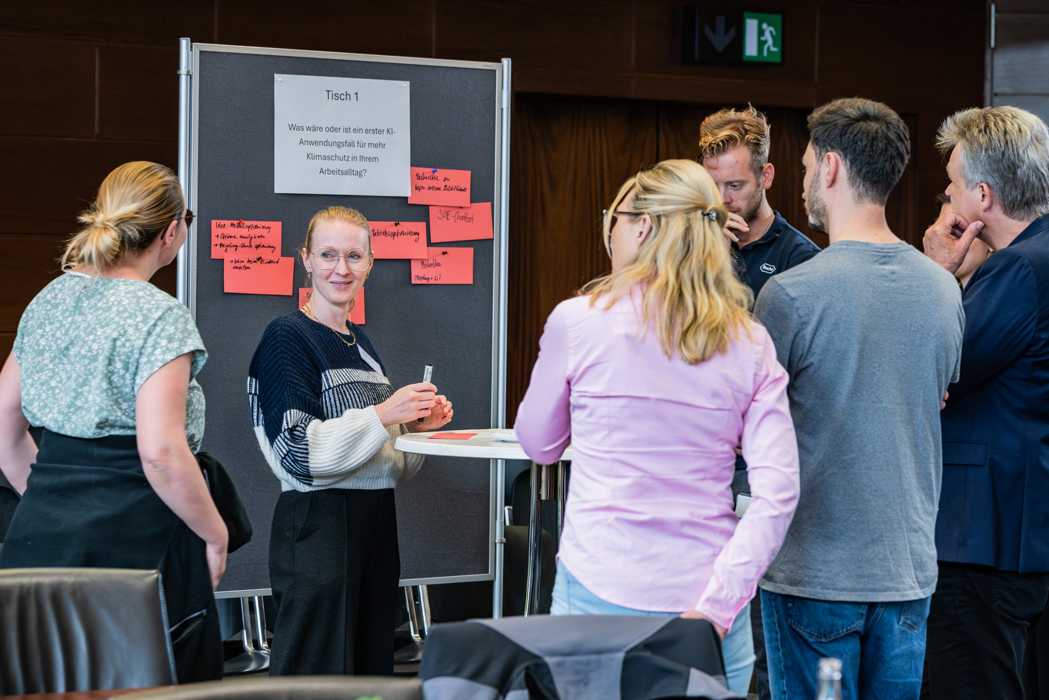 Eine Gruppe von Menschen versammelt sich um einen Stehtisch, an dem eine Frau steht und Informationen präsentiert. Im Hintergrund befinden sich Pinnwände mit pinken Notizzetteln. Die Atmosphäre ist interaktiv und kommunikativ, mit einem Fokus auf den Austausch von Ideen.