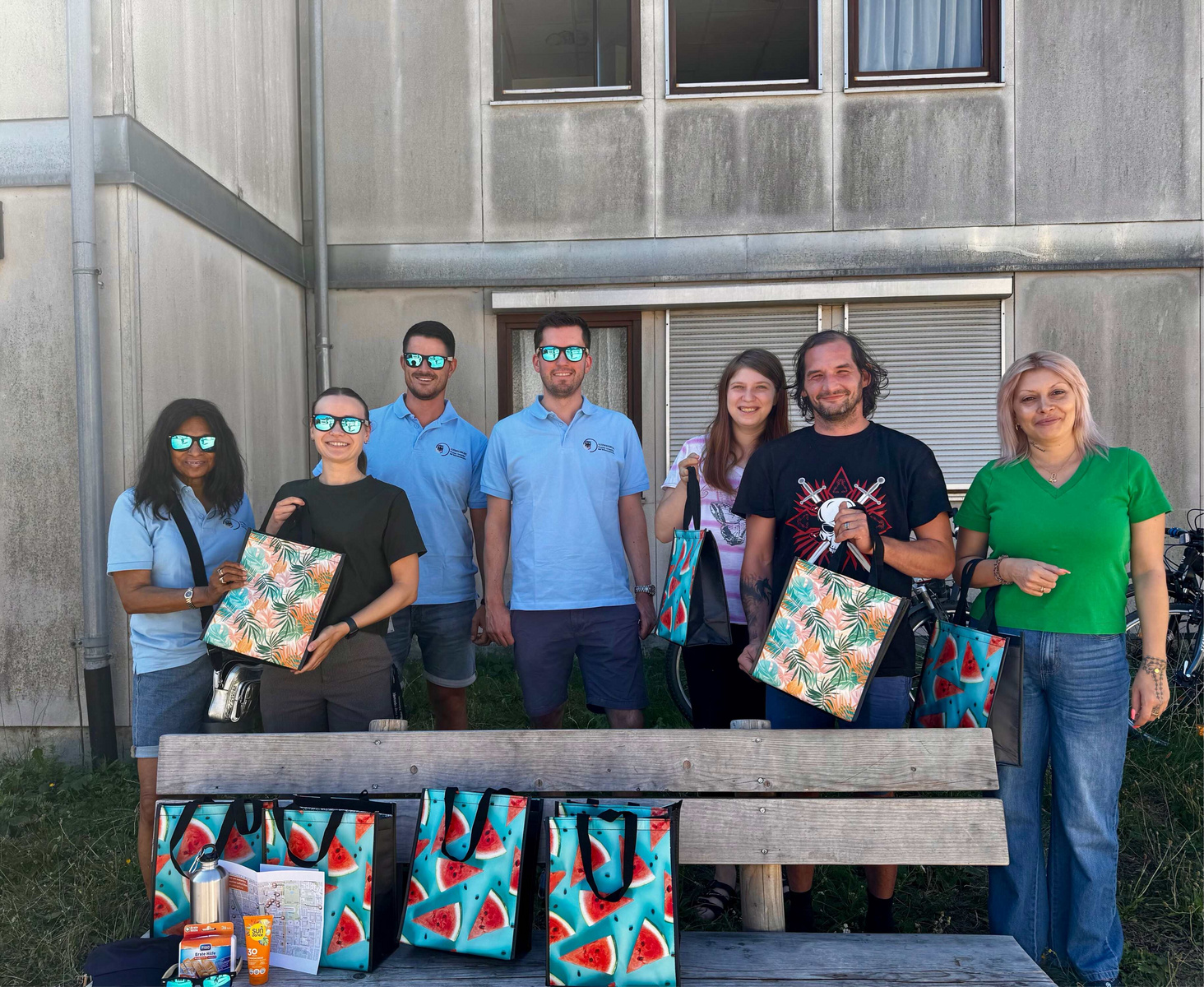 Eine Gruppe von acht Personen steht vor einem Gebäude und hält farbenfrohe Taschen mit Wassermelonen-Motiv. Alle tragen Sonnenbrillen und lächeln. Im Vordergrund sind einige gesunde Snacks auf einer Bank sichtbar.