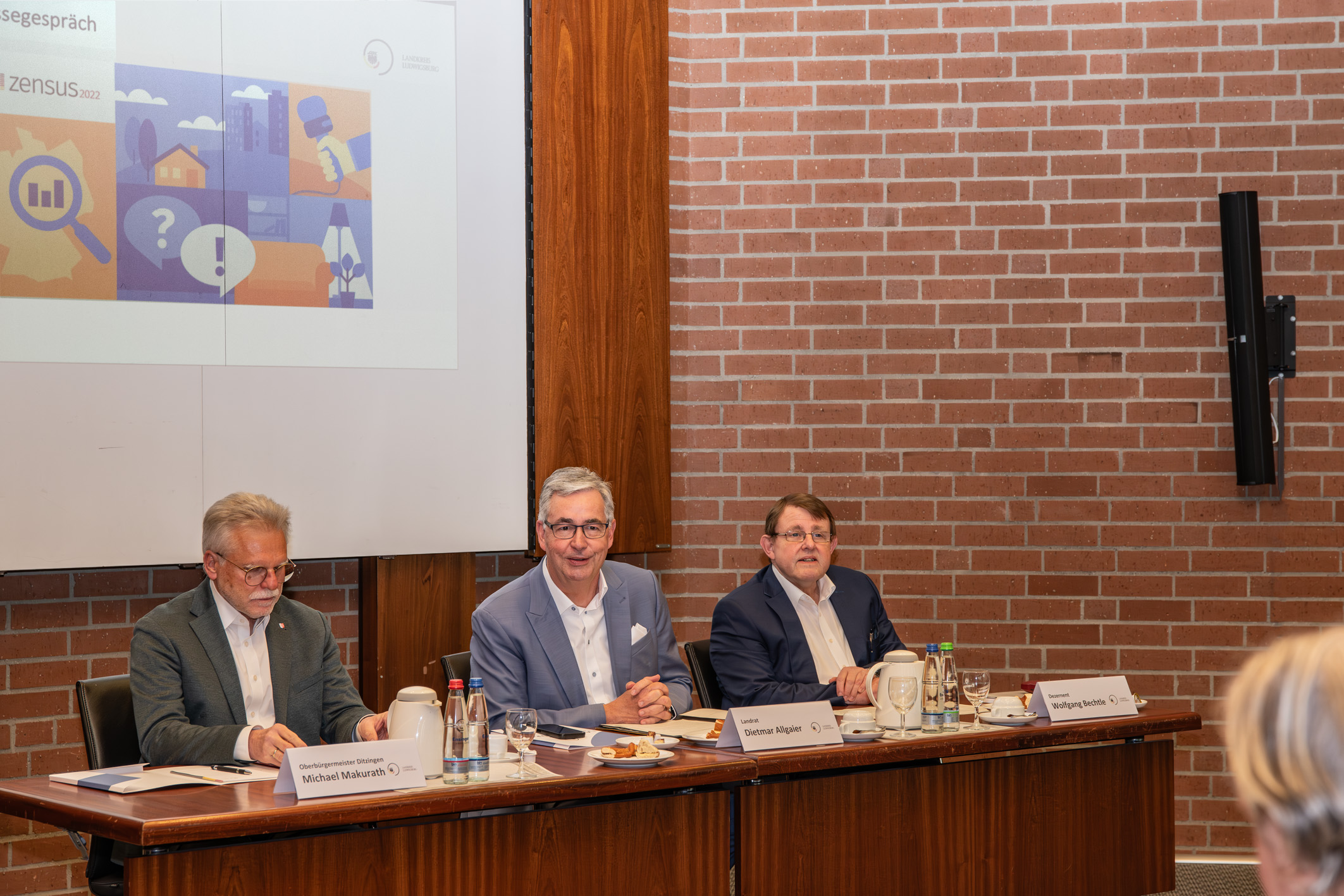 Drei Männer sitzen an einem Tisch bei einer Veranstaltung. Im Hintergrund ist eine Präsentation mit Grafiken und Diagrammen zu sehen. Die Männer scheinen über ein wichtiges Thema zu sprechen, während auf dem Tisch Getränke und Notizen liegen.