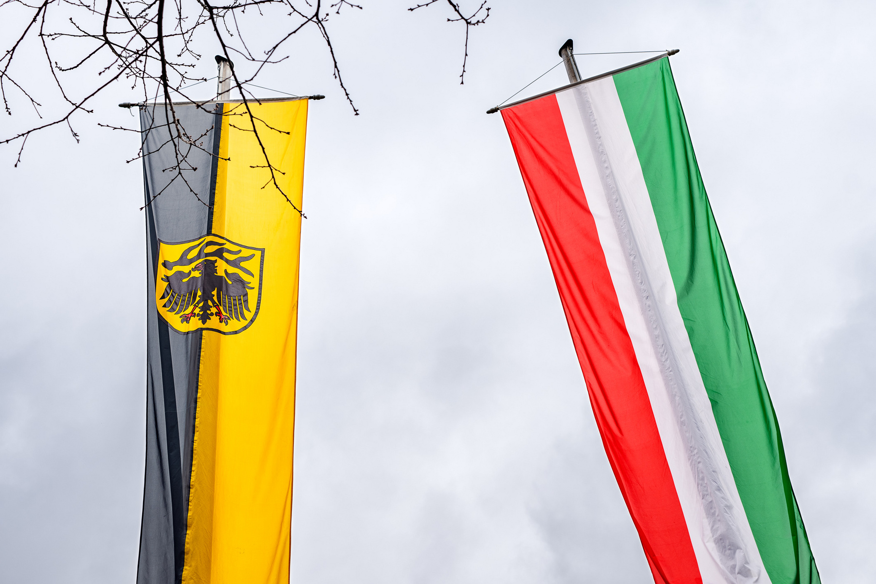 Zwei Fahnen wehen im Wind. Links die deutsche Flagge in Schwarz-Gelb mit dem Bundesadler, rechts die italienische Flagge in Grün, Weiß und Rot. Der Himmel ist bewölkt.