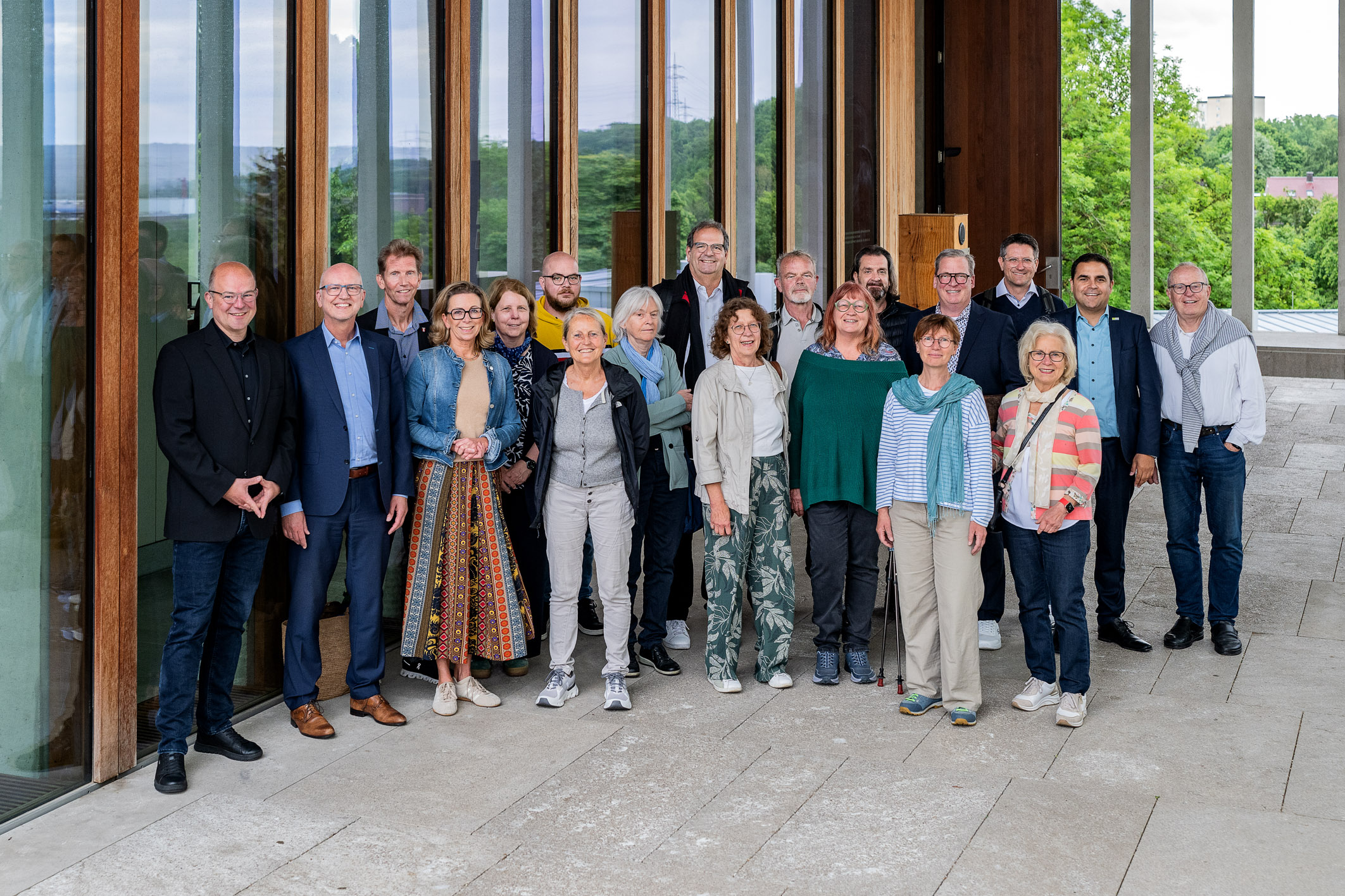 Eine Gruppe von etwa 20 Personen steht im Freien vor einem Gebäude mit großen Fenstern. Die Teilnehmer sind unterschiedlich gekleidet und lächeln. Die Umgebung ist grün und die Atmosphäre wirkt freundlich und einladend.