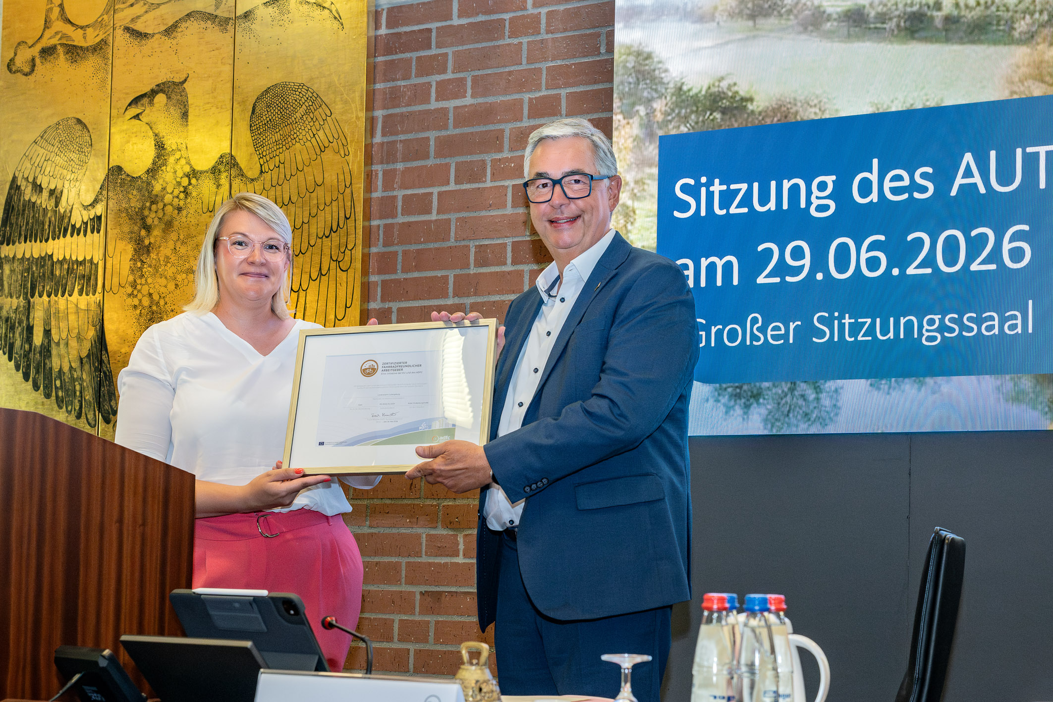 Die Abbildung zeigt eine Frau und einen Mann, die sich in einem Sitzungssaal gegenüberstehen. Der Mann überreicht der Frau ein Zertifikat oder eine Auszeichnung. Im Hintergrund ist ein Banner mit dem Datum und dem Titel der Sitzung zu sehen.